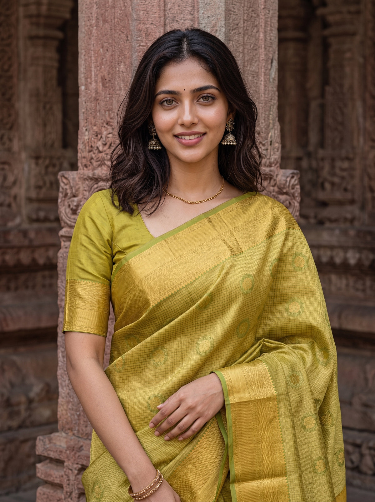 A woman wearing a yellow-green silk saree with a matching short-sleeve blouse, standing in front of a carved stone pillar.