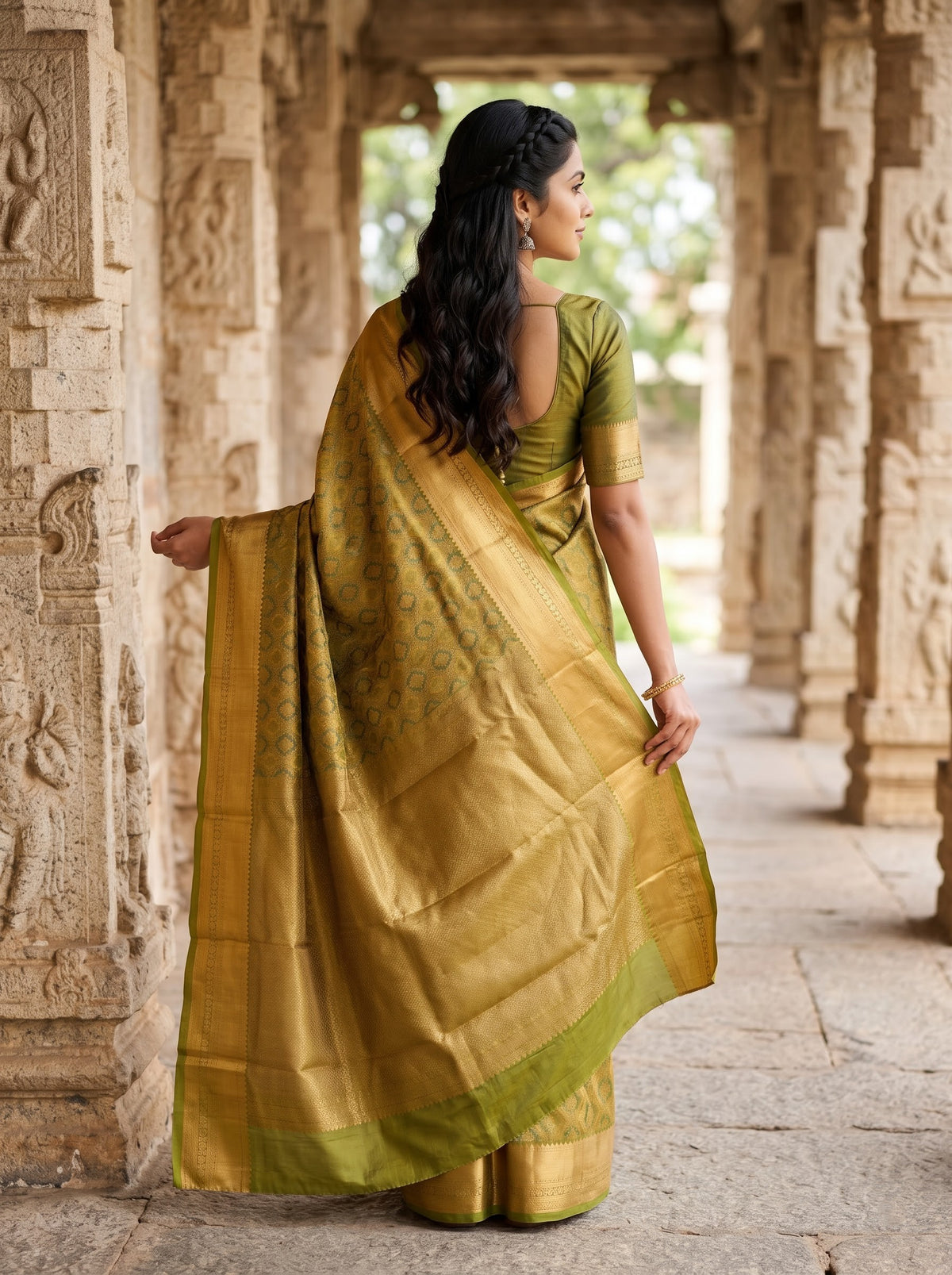 A woman wearing a golden-yellow and olive green silk saree, standing in a stone corridor with carved pillars.