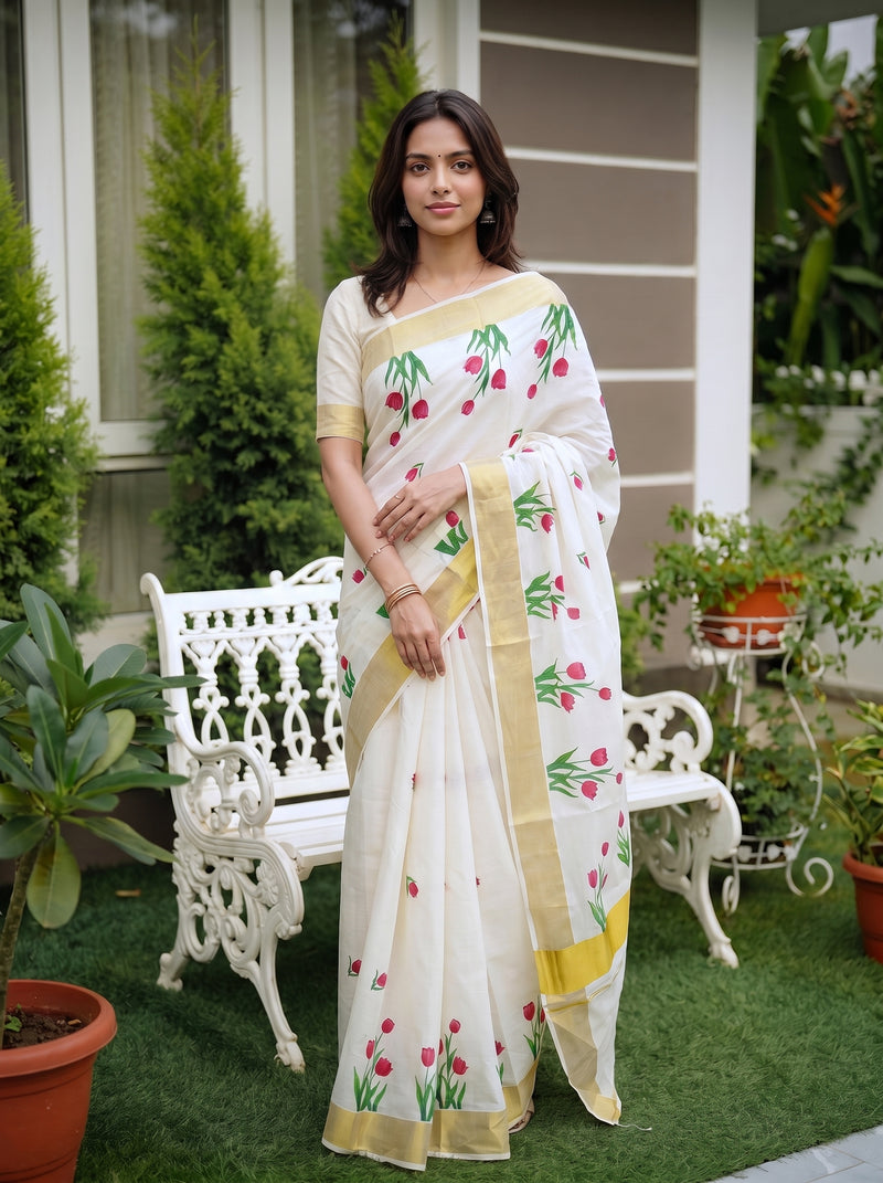 A woman wearing a white saree with pink tulip prints and gold borders, standing in a garden courtyard next to a white bench.