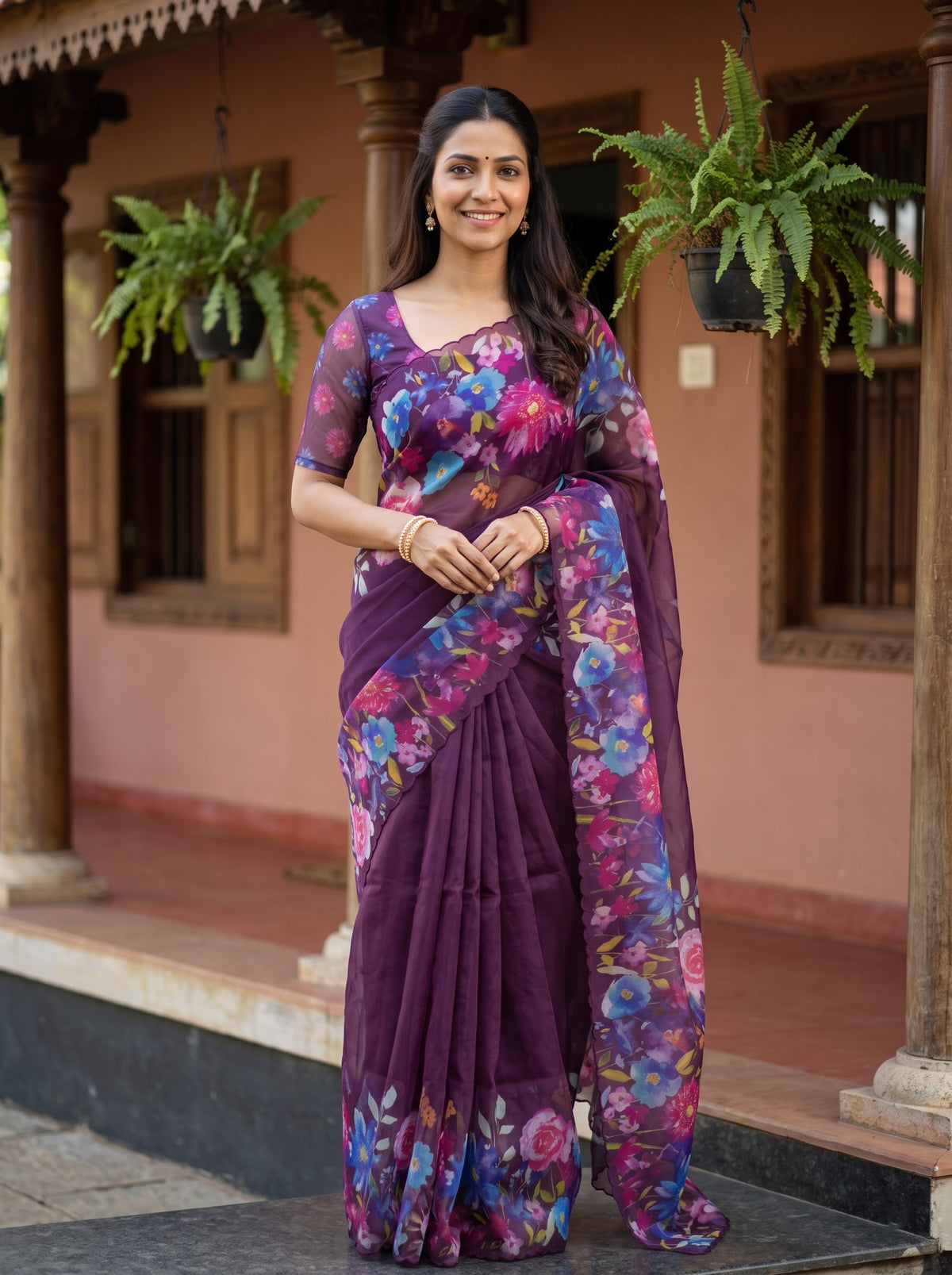 A woman wearing a purple floral printed saree stands on the steps of a traditional building with pink walls and hanging green plants.
