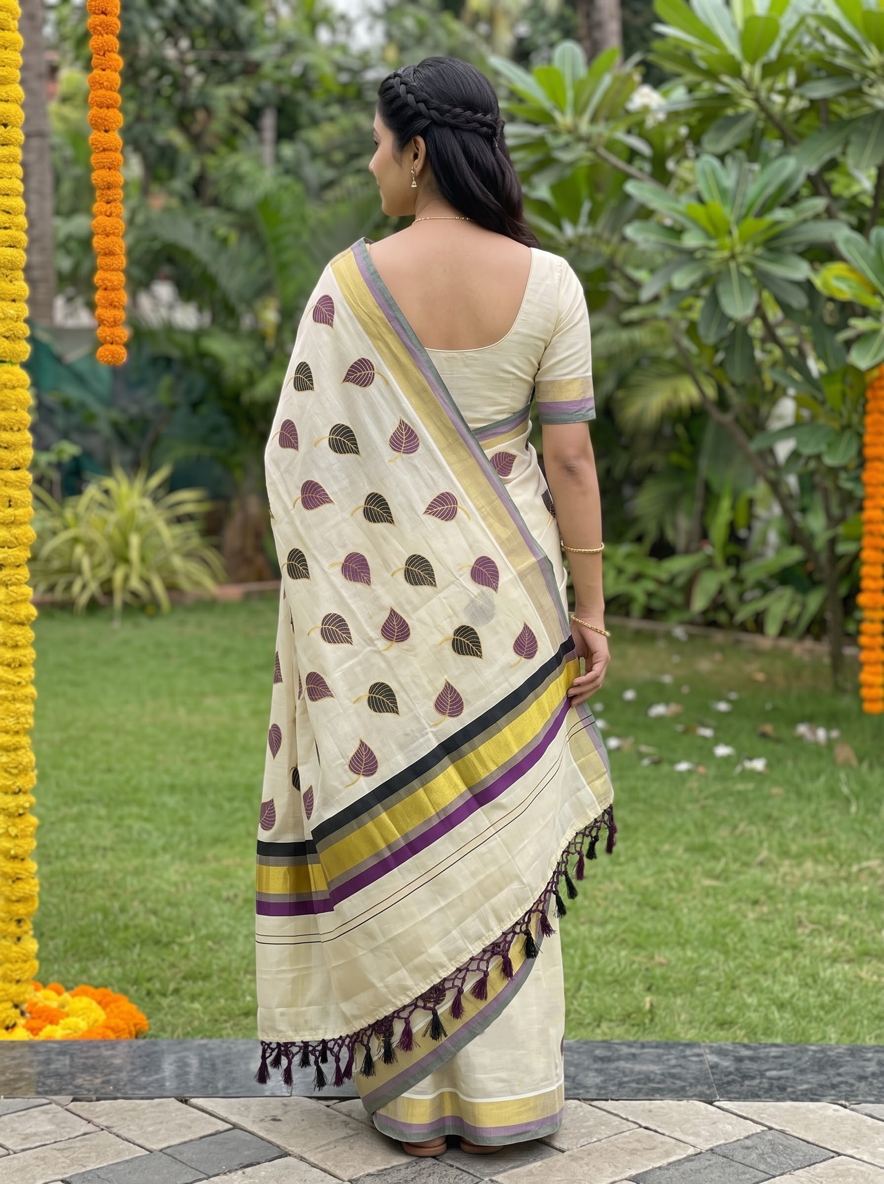 A woman stands outdoors in a garden wearing a cream-colored saree with purple and gold printed motifs, a purple and gold border, and a matching cream blouse. She is shown from the back, with green foliage and hanging orange marigold garlands in the background.