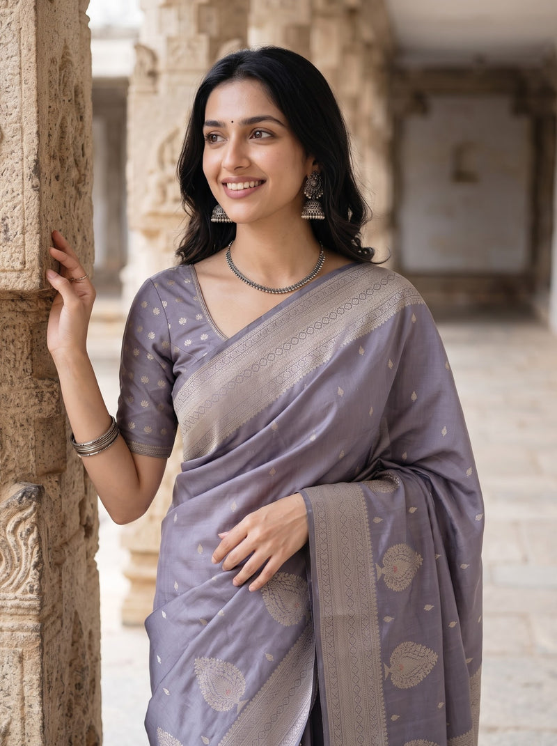 A woman wearing a lavender-gray silk saree with gold zari motifs and a matching blouse, standing in a stone corridor next to a carved pillar.