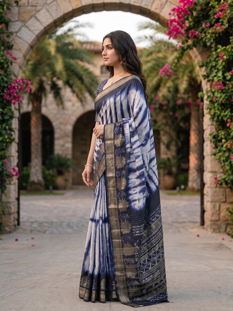 A woman wearing a blue and white tie-dye saree with a patterned border, standing in a stone archway corridor with palm trees and pink flowers in the background.