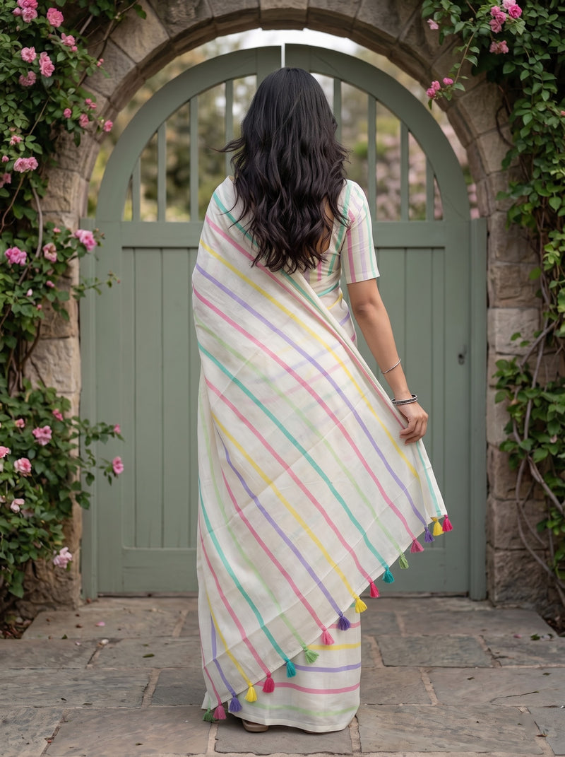 A woman viewed from behind wearing a white saree with pastel pink, green, yellow, and blue stripes and colorful tassels, standing in front of a stone archway with a green door and climbing pink flowers.