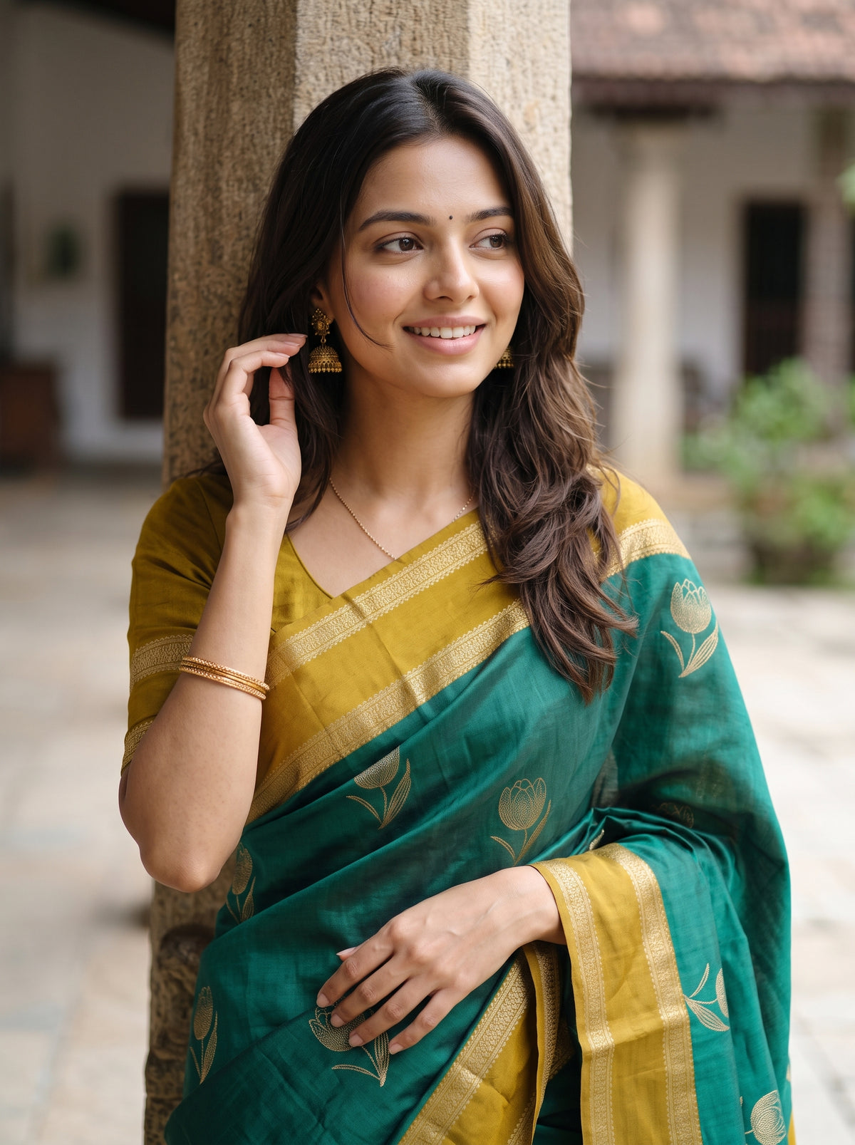 A woman wearing a green and mustard yellow saree with gold floral motifs, standing outdoors against a stone pillar.