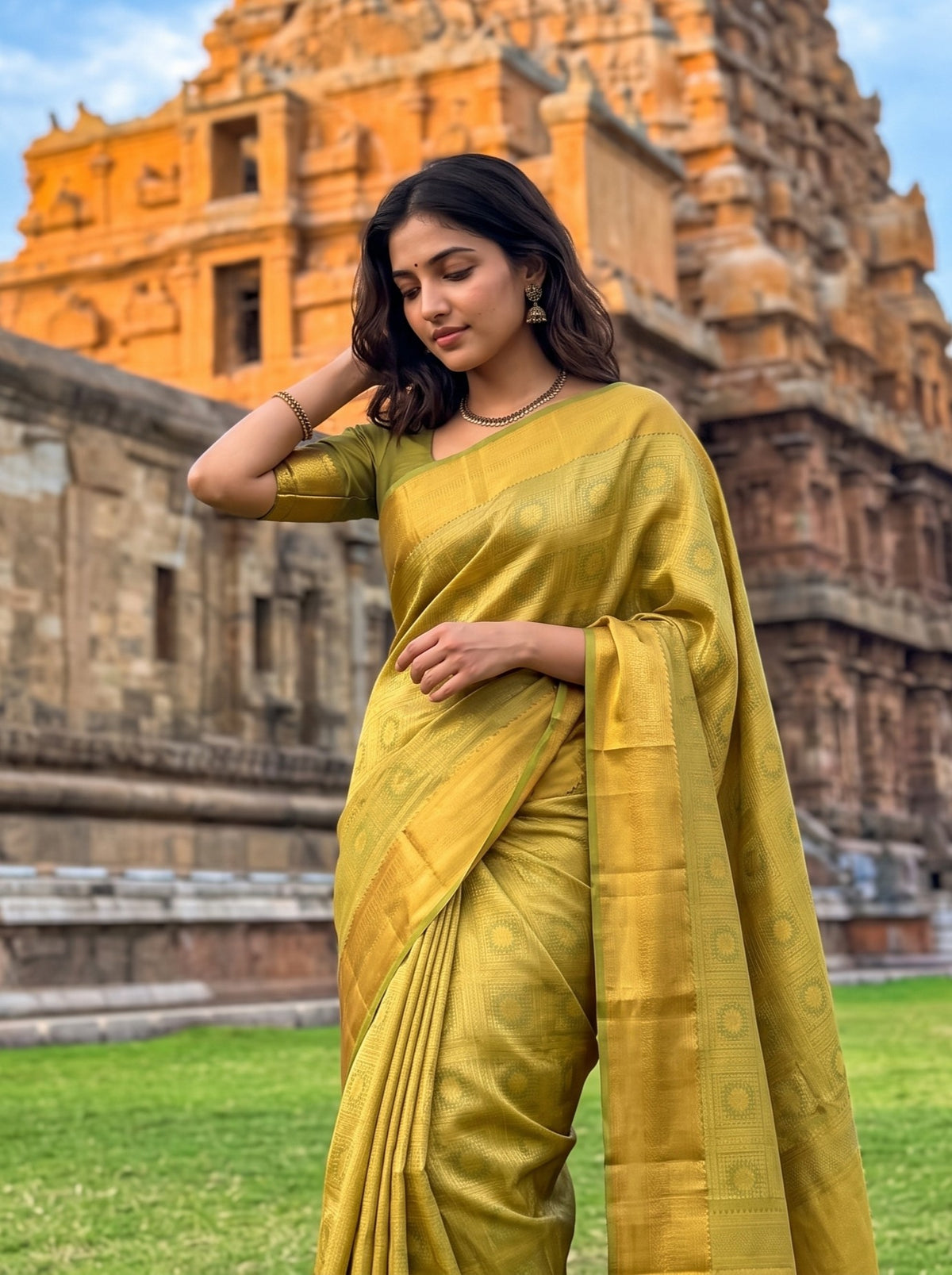 A woman wearing a golden yellow silk saree with a matching short-sleeve blouse stands outdoors in front of a historic temple structure and green grass.