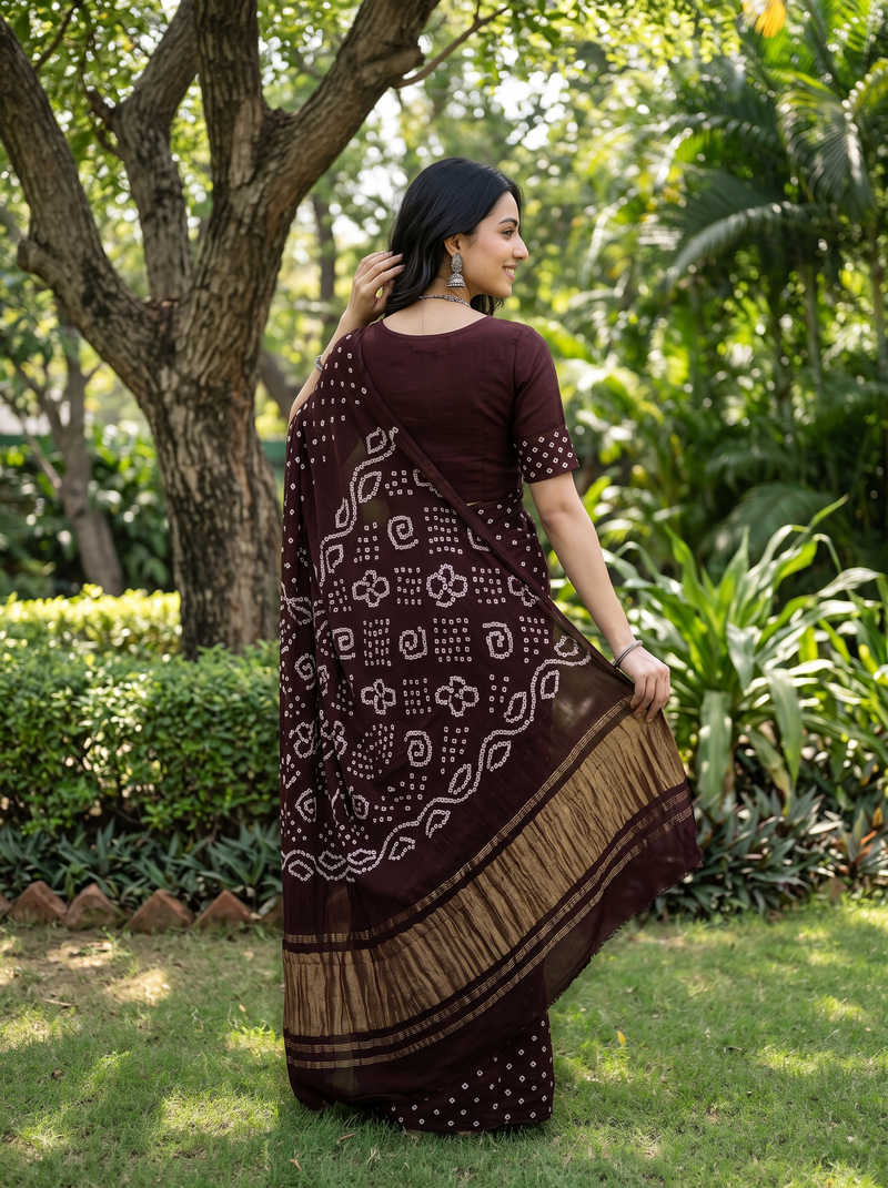 A woman wearing a dark brown and beige printed saree stands outdoors in a garden, holding out the fabric to show its pattern and border.