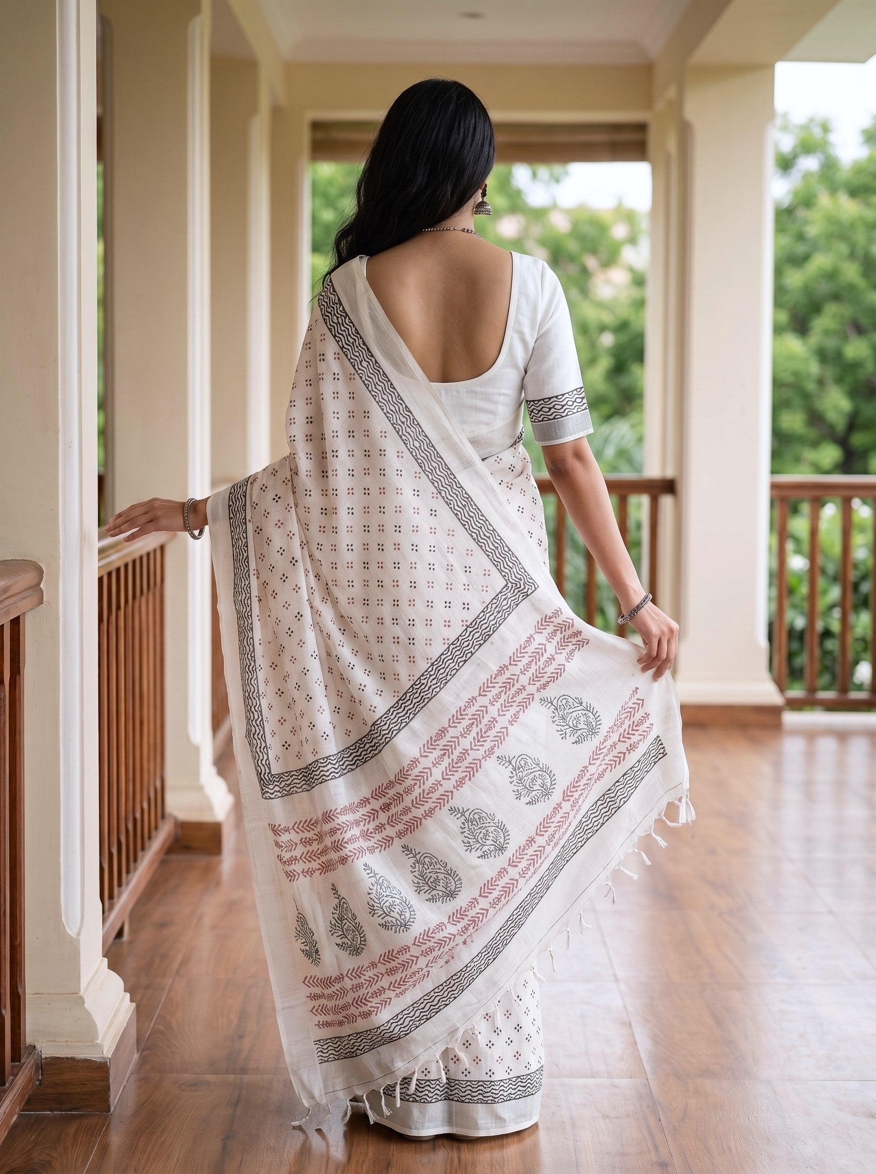 A woman viewed from behind wearing a white saree with black and pink printed borders and motifs, standing on a wooden-floored indoor porch with columns and greenery outside.
