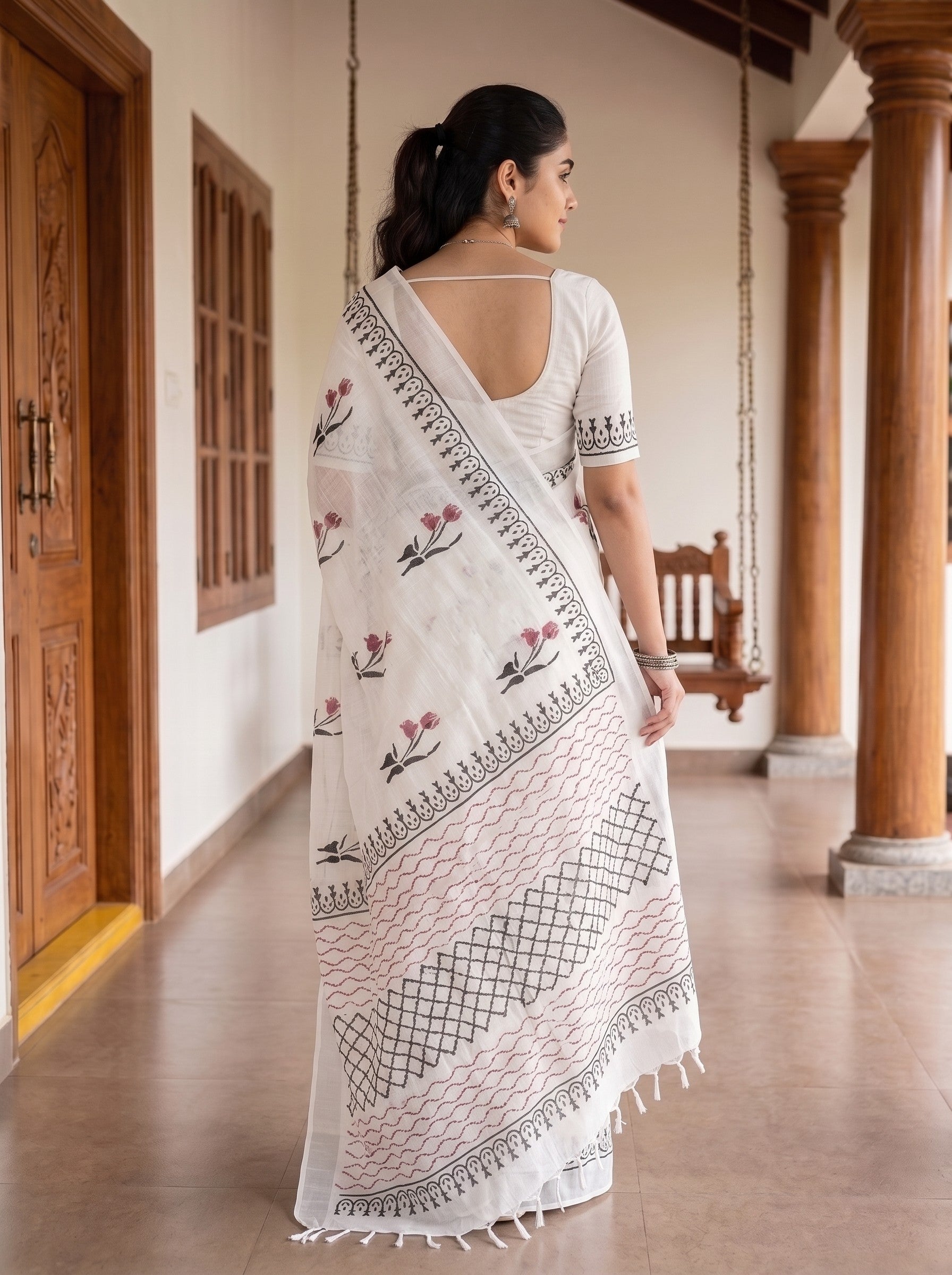 A woman stands in a corridor wearing a white saree with black geometric border patterns and pink floral motifs, paired with a white blouse.