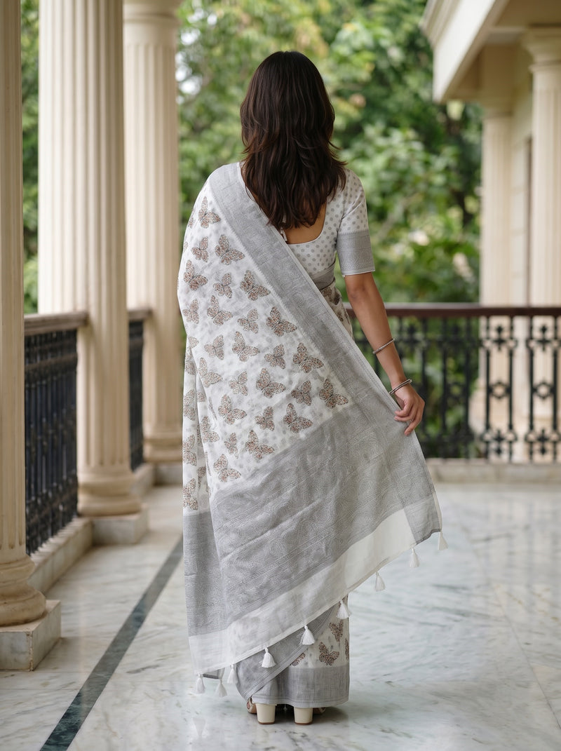 A woman viewed from behind wearing a light gray and white saree with brown floral prints, standing on a marble balcony with columns and green foliage in the background.
