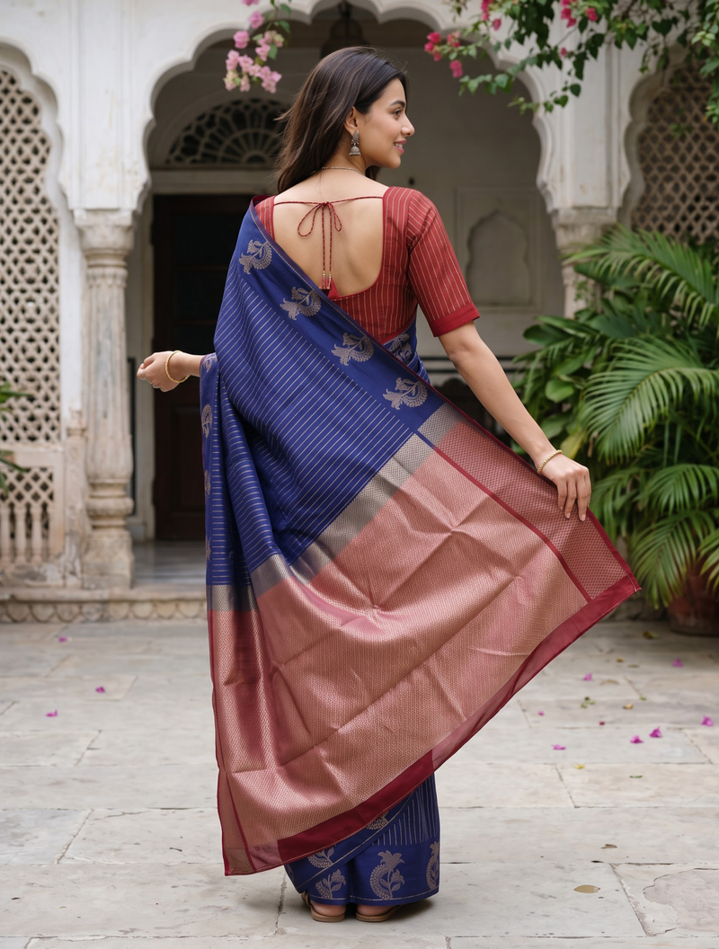 A woman wearing a blue and rose gold silk saree with a red blouse, photographed from behind in an outdoor courtyard setting.