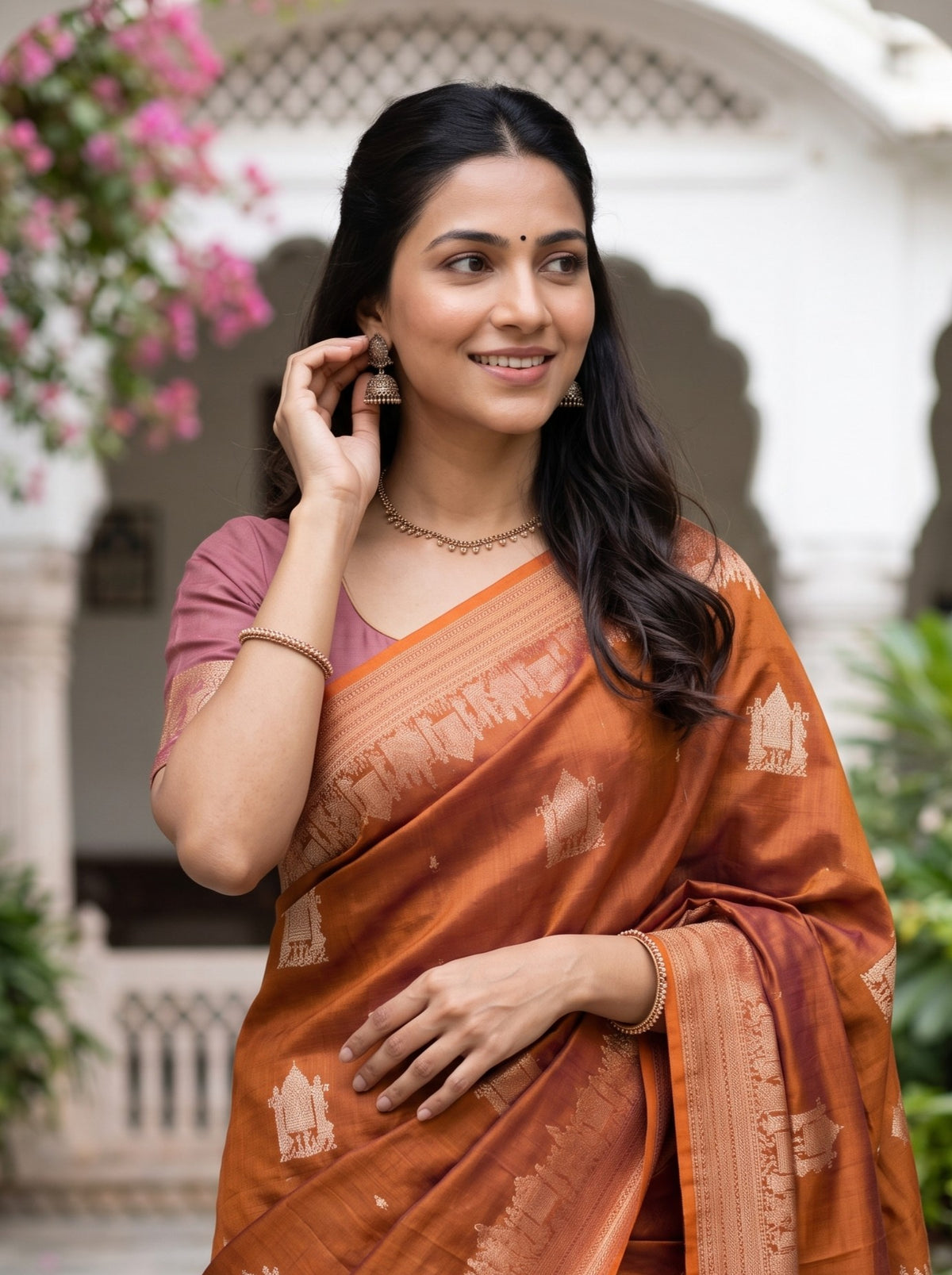 A woman wearing a rust-orange and brown patterned silk saree with a dusty pink blouse, standing in front of a white arched building with pink flowers.