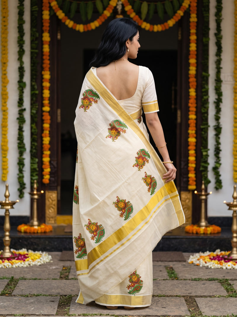 A woman wearing a cream-colored cotton saree with yellow borders and green floral motifs, standing in front of a decorated doorway with marigold garlands.