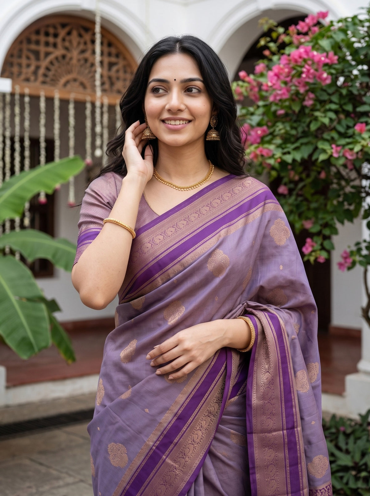 A woman wearing a lavender purple silk saree with gold zari patterns and borders, standing in front of a traditional courtyard with pink flowers.
