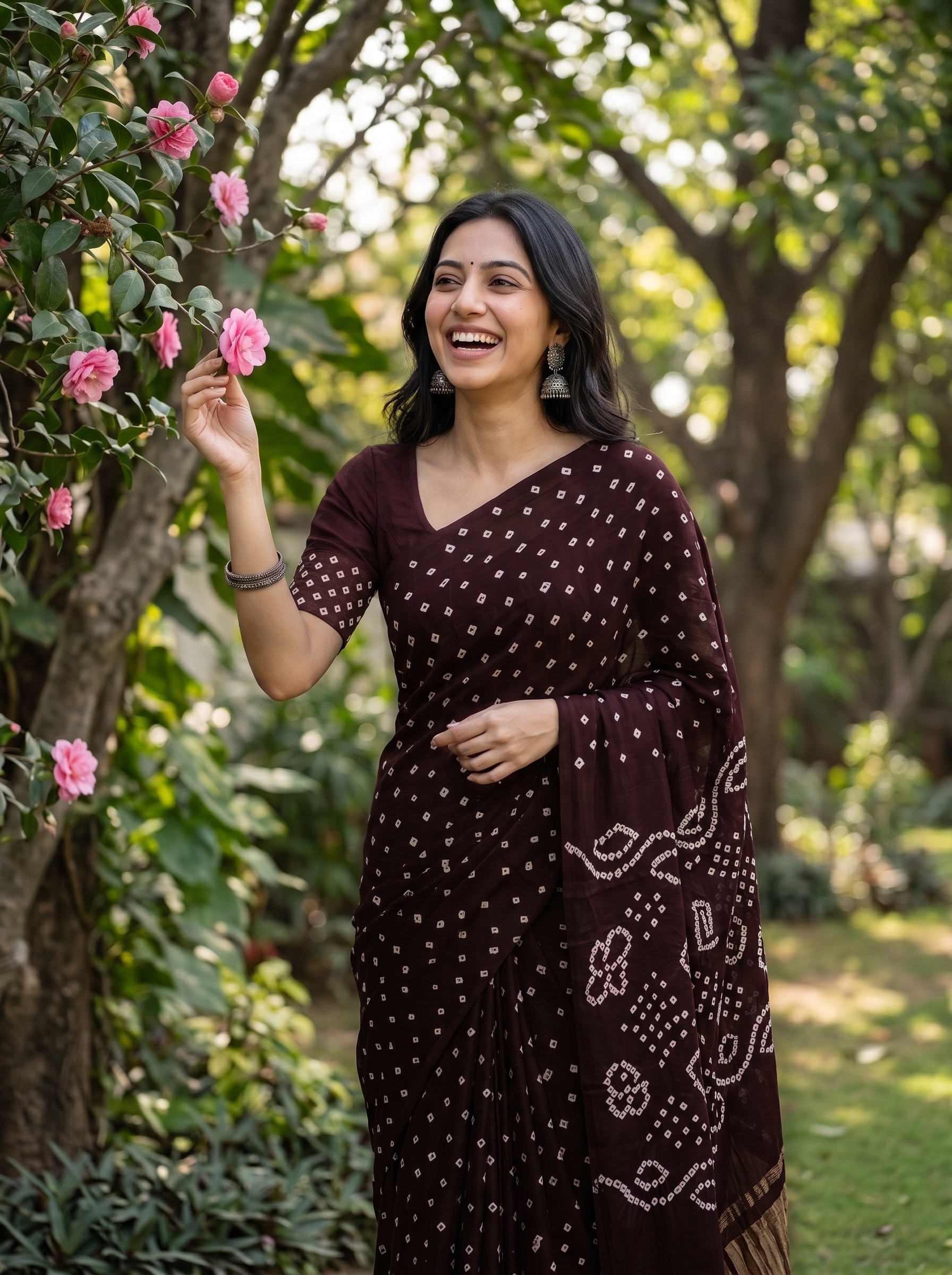 A woman wearing a dark brown Bandhani saree with white tie-dye patterns stands in a garden, smiling and touching pink flowers on a tree.