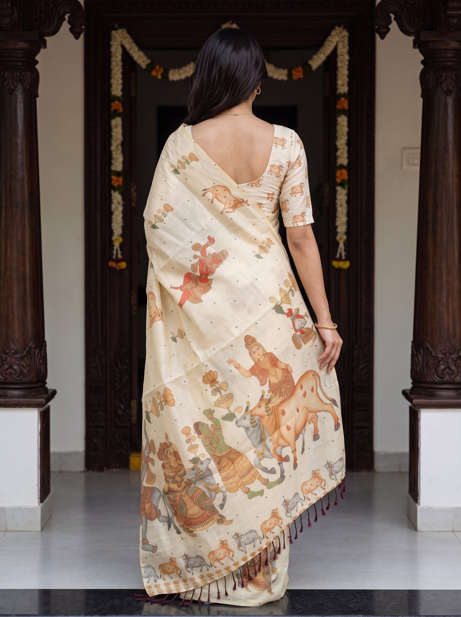 A woman viewed from behind wearing a cream-colored cotton saree with a matching short-sleeve blouse. The saree features a detailed, multicolor traditional Indian illustration pattern depicting figures and animals.