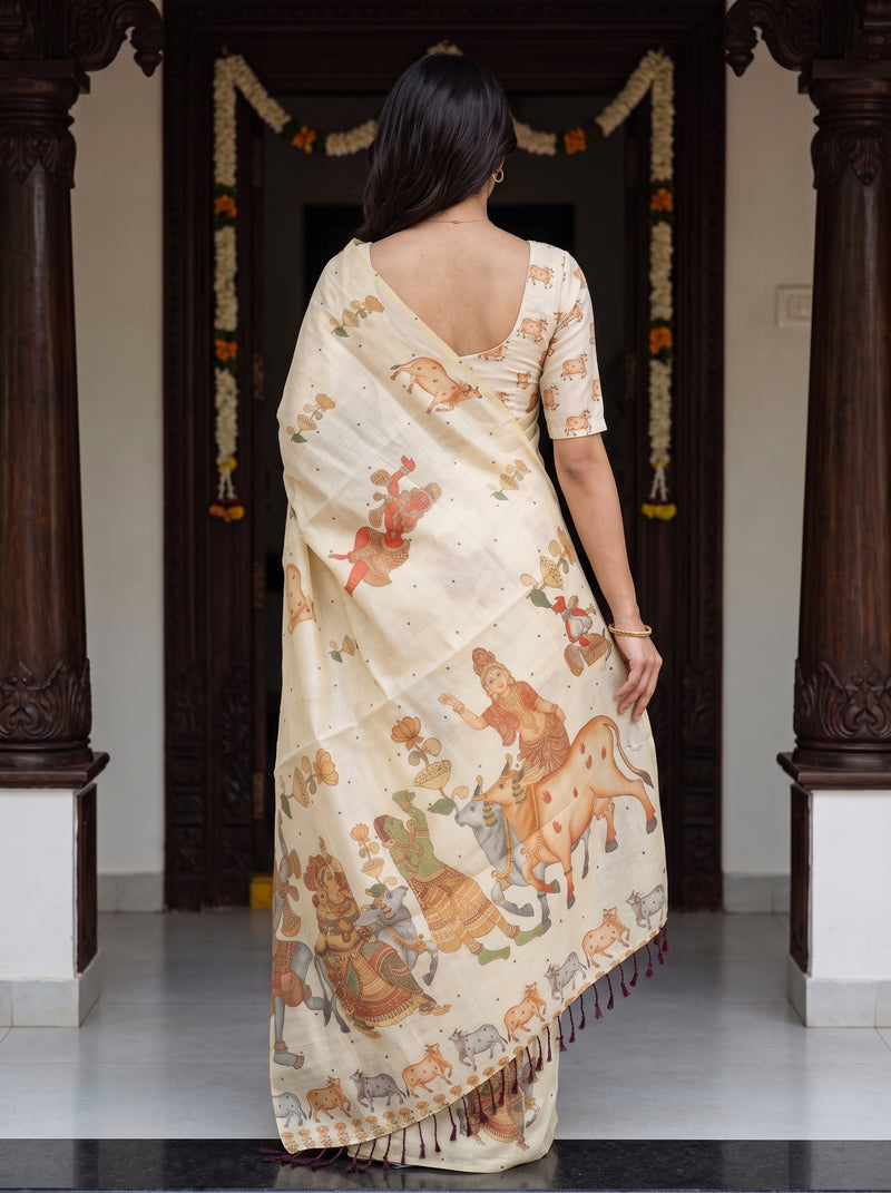 A woman viewed from behind wearing a cream-colored cotton saree with a matching short-sleeve blouse. The saree features a detailed, multicolor traditional Indian illustration pattern depicting figures and animals.