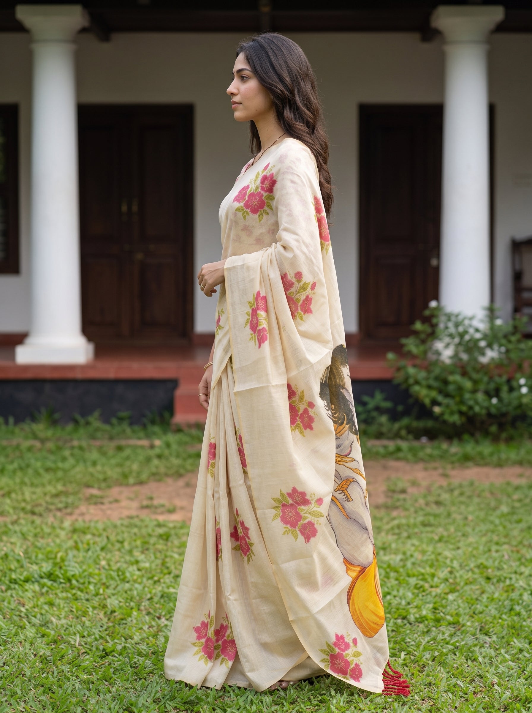 A woman wearing a cream-colored saree with pink floral prints and a yellow border, standing outdoors on a green lawn in front of a building with white pillars.