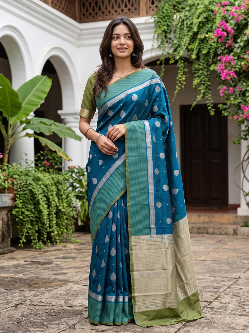 A woman wearing a teal blue and green saree with silver motifs and a beige border, standing in a courtyard with white walls and green plants.