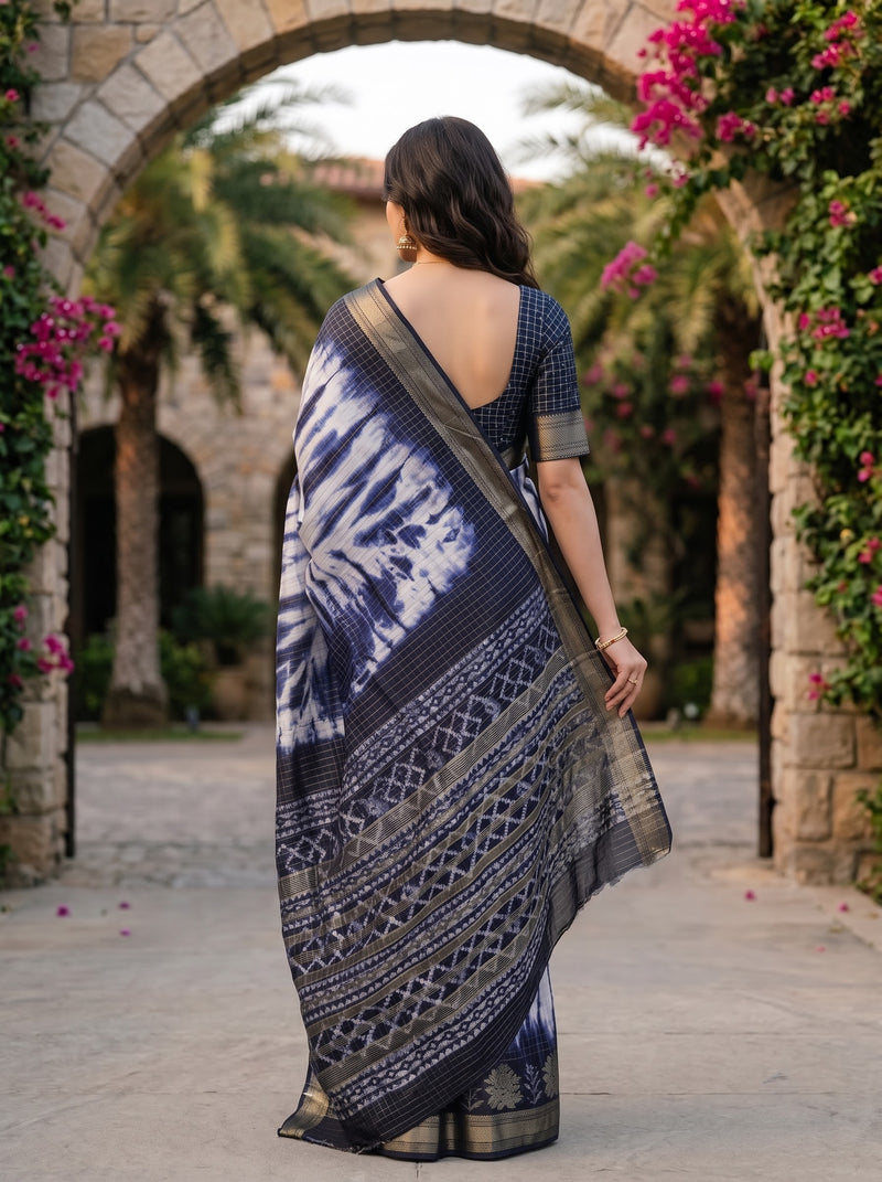 A woman viewed from behind wearing a navy blue and white patterned saree with a dark blue patterned blouse, standing in front of a stone archway decorated with pink flowers and palm trees.