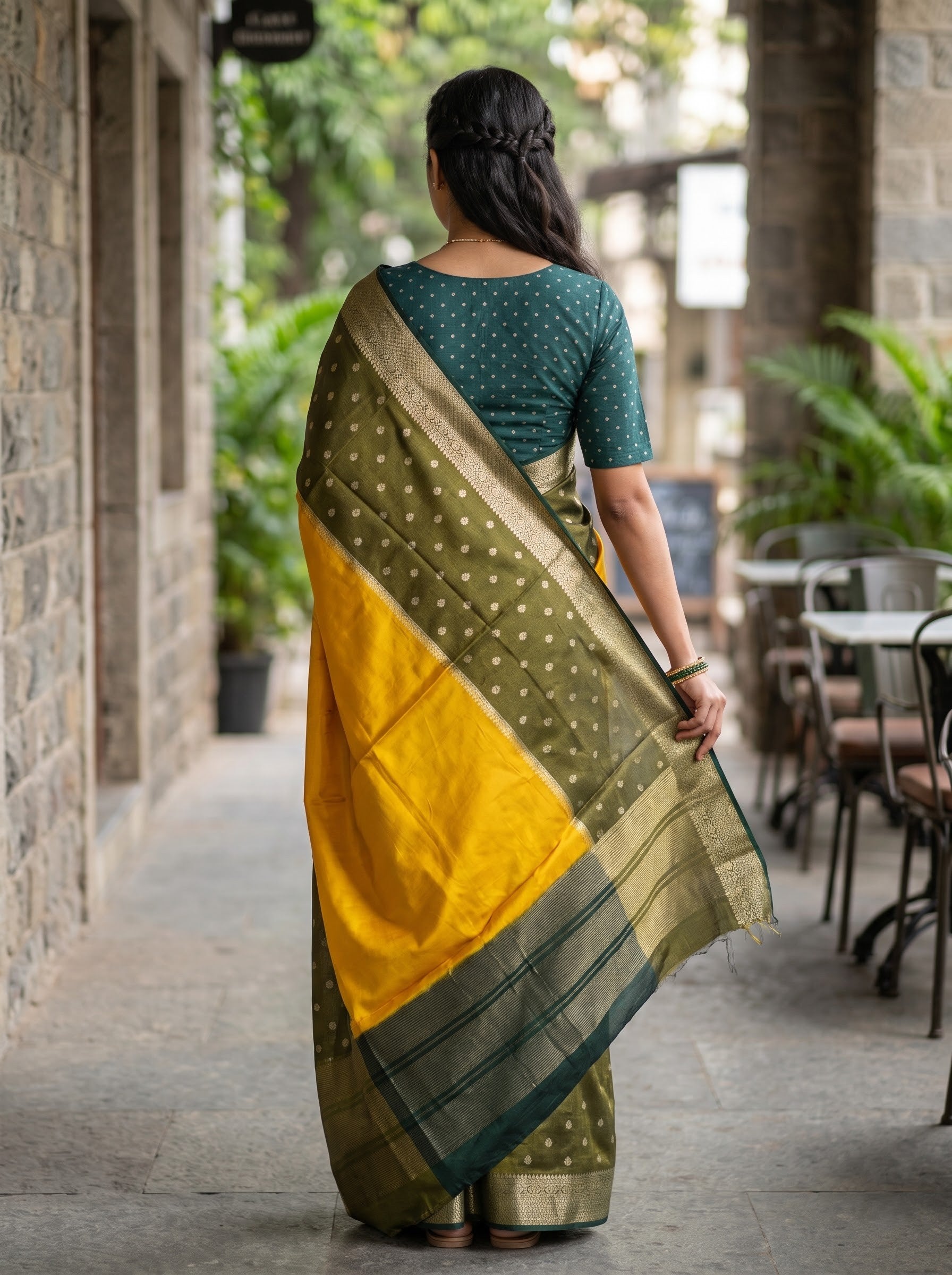 A woman viewed from behind wearing a mustard yellow and olive green silk saree with a dark teal polka dot blouse, standing in a stone corridor.