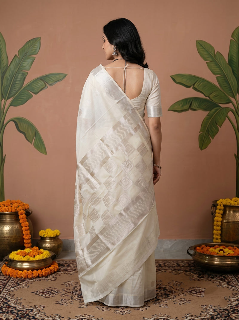 A woman stands with her back to the camera wearing a white saree with a matching blouse. The setting features a terracotta-colored wall, decorative potted banana plants, and brass pots with orange marigold garlands on a patterned rug.