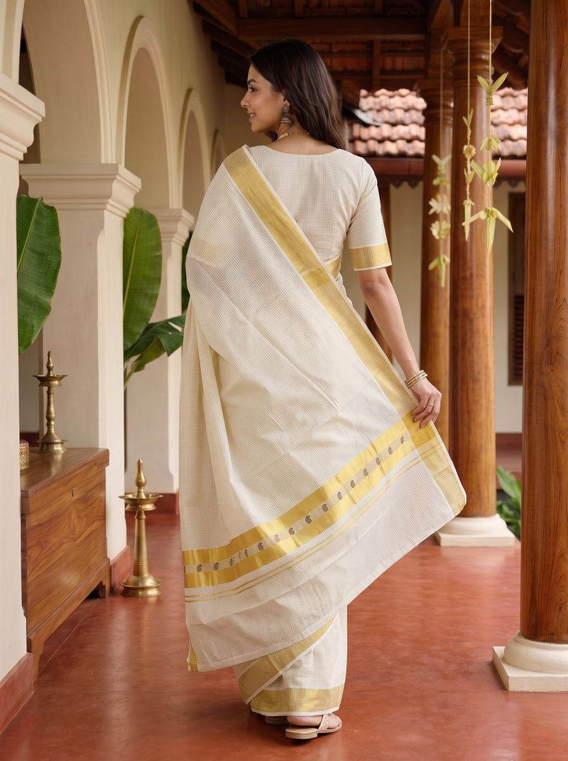 A woman wearing a cream-colored cotton saree with a gold border and a matching cream blouse, standing in a traditional courtyard with wooden pillars.