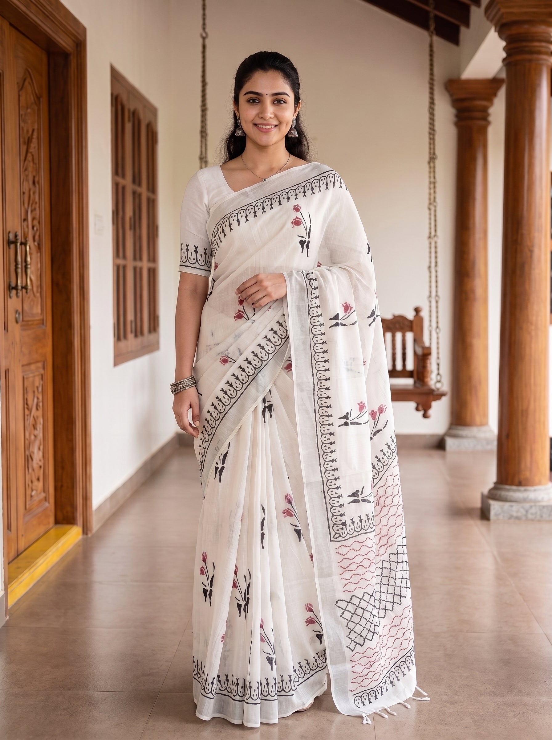 A woman wearing a white saree with black and pink floral embroidery, standing in a traditional corridor with wooden pillars.