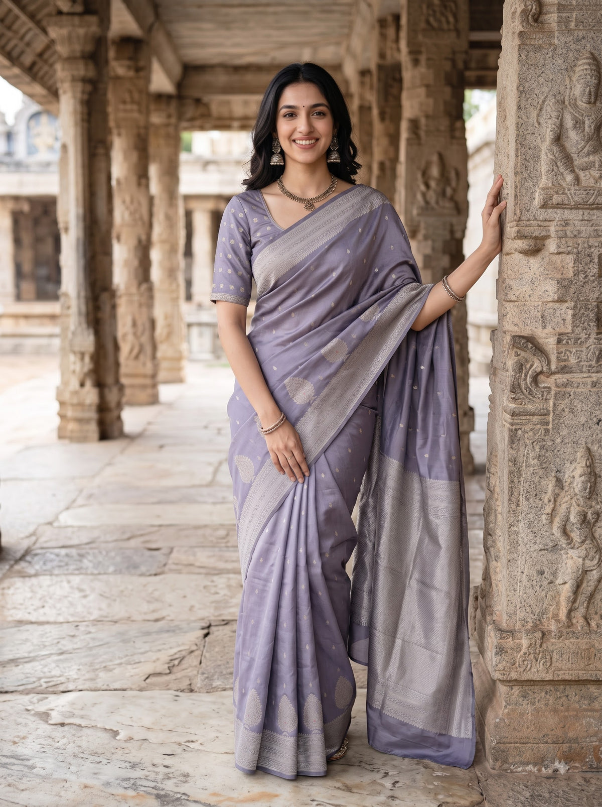A woman wearing a lavender and gray patterned saree stands in a stone corridor with carved pillars, posing and holding a stone column.