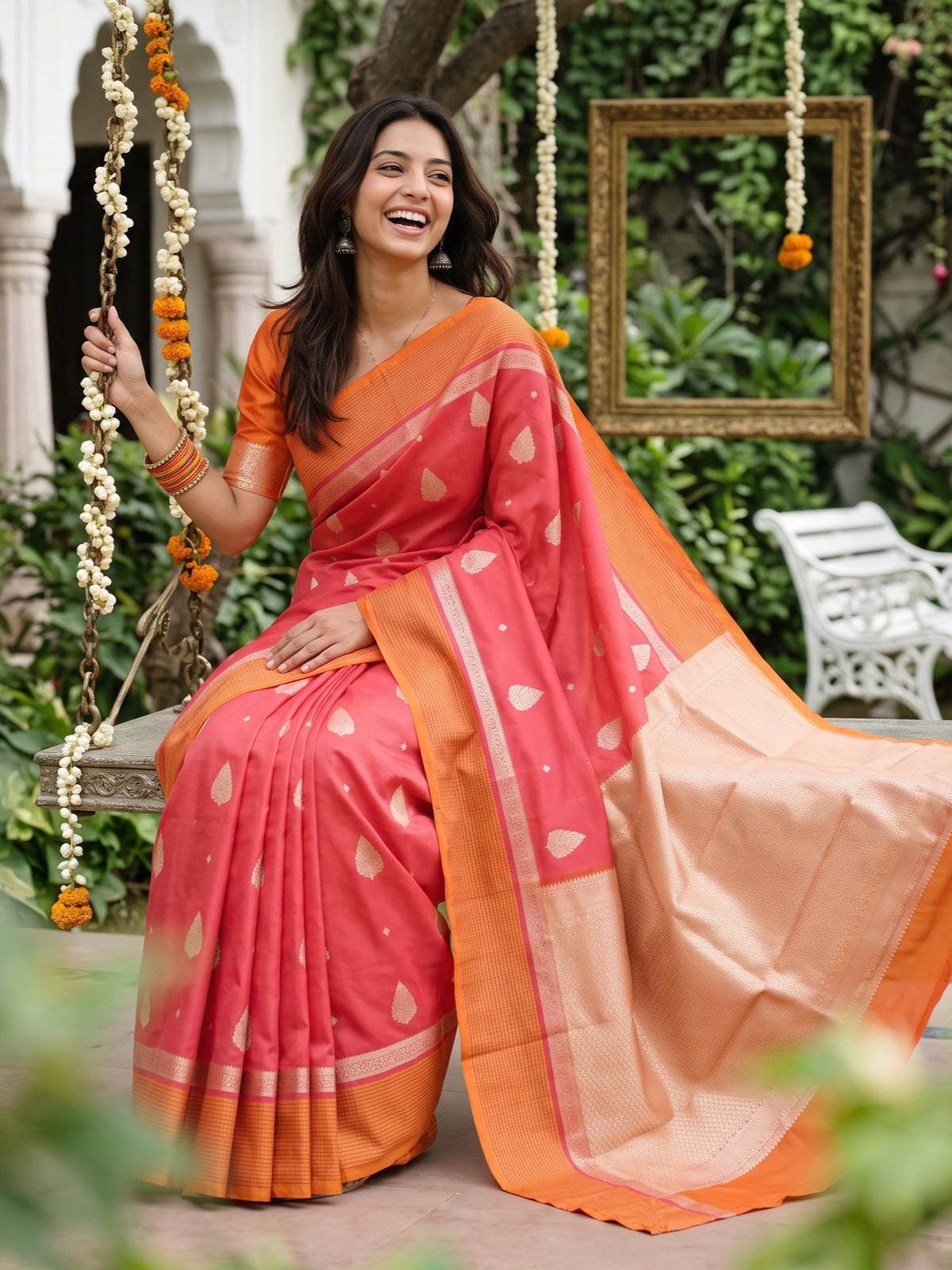 A woman seated on a hanging swing in a garden courtyard wearing a coral pink and orange saree with peach border details. The scene includes greenery, a mirror, and decorative garlands.