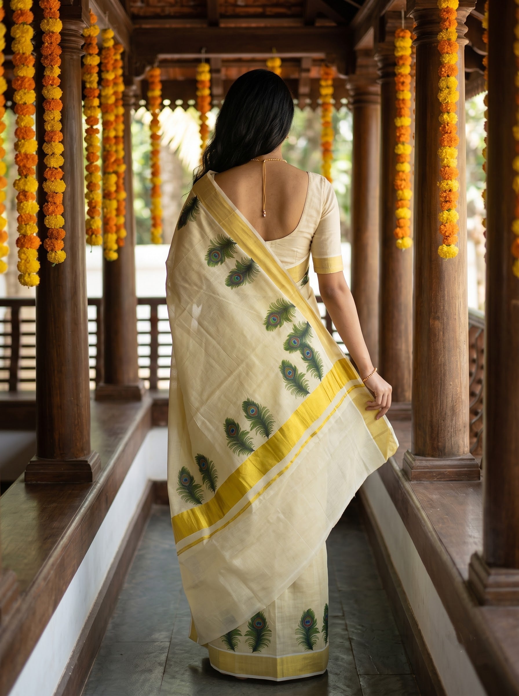 A woman viewed from behind wearing a cream-colored saree with green leaf prints and a yellow border, standing in a traditional wooden corridor decorated with orange and yellow marigold garlands.