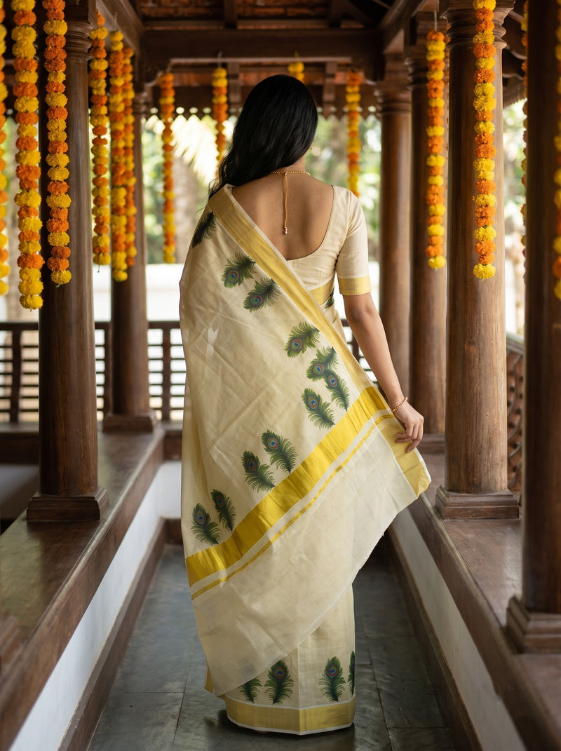 A woman viewed from behind wearing a cream-colored saree with green leaf prints and a yellow border, standing in a traditional wooden corridor decorated with orange and yellow marigold garlands.
