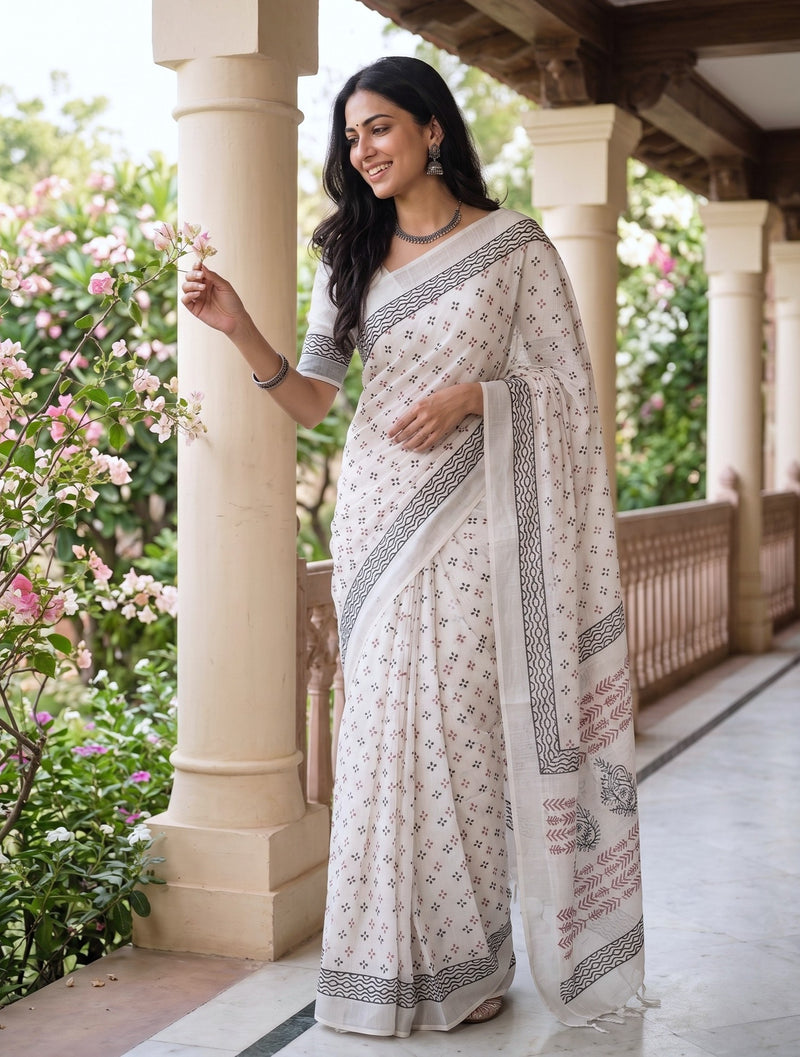 A woman wearing a white printed saree with black and pink border patterns, standing on a traditional stone porch with beige pillars and green foliage in the background.