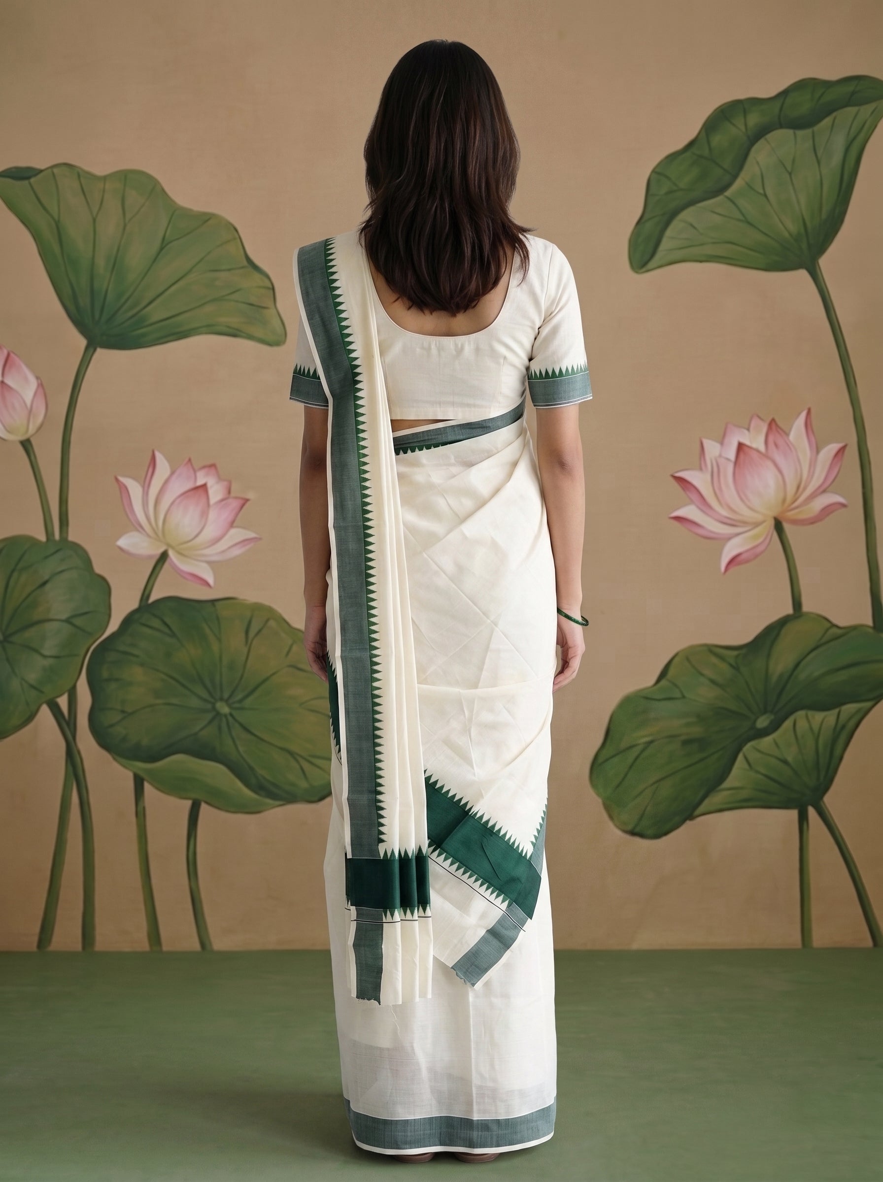 A woman stands with her back to the camera wearing a white saree with dark green border accents and a matching blouse, posed in front of a painted backdrop featuring lotus leaves and pink lotus flowers.