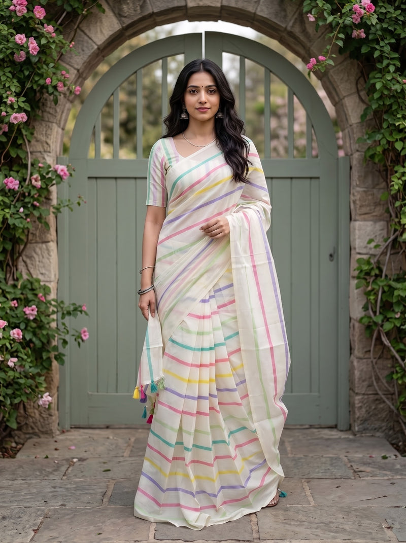 A woman wearing a white cotton saree with pastel rainbow stripes stands in front of a stone archway with climbing pink roses.
