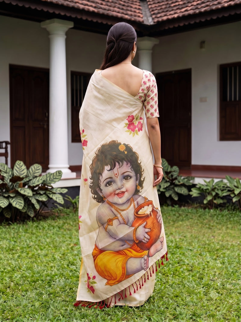 A woman stands outdoors in a garden wearing a cream-colored saree with a large printed illustration of a child and pink floral accents. The background shows a house with white pillars, dark brown doors, and green grass.
