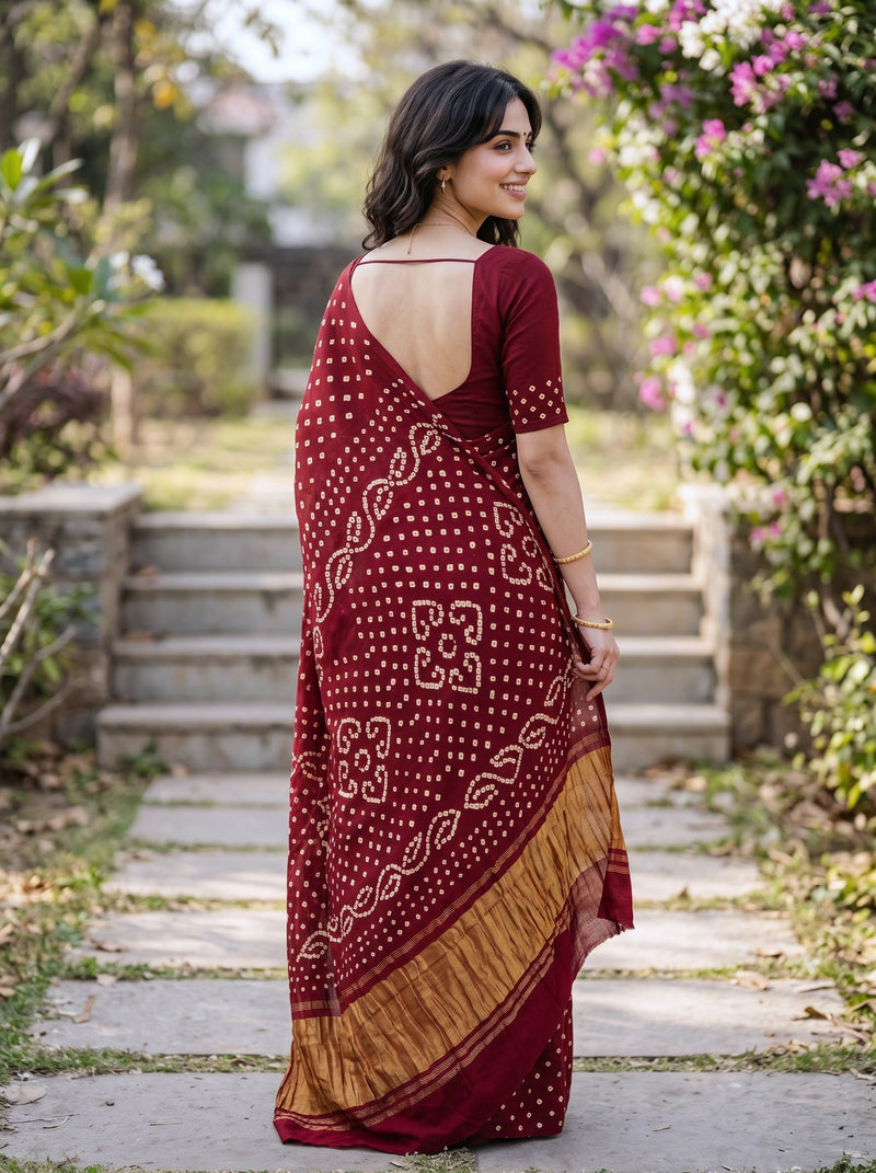 A woman wearing a maroon and gold printed saree, photographed from behind in an outdoor garden setting with stone steps and greenery.