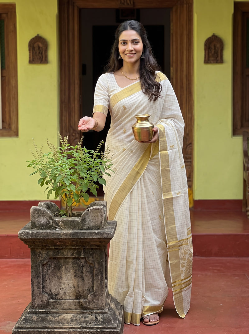 A woman wearing a cream and gold checkered saree stands in a courtyard holding a brass pot next to a potted plant.