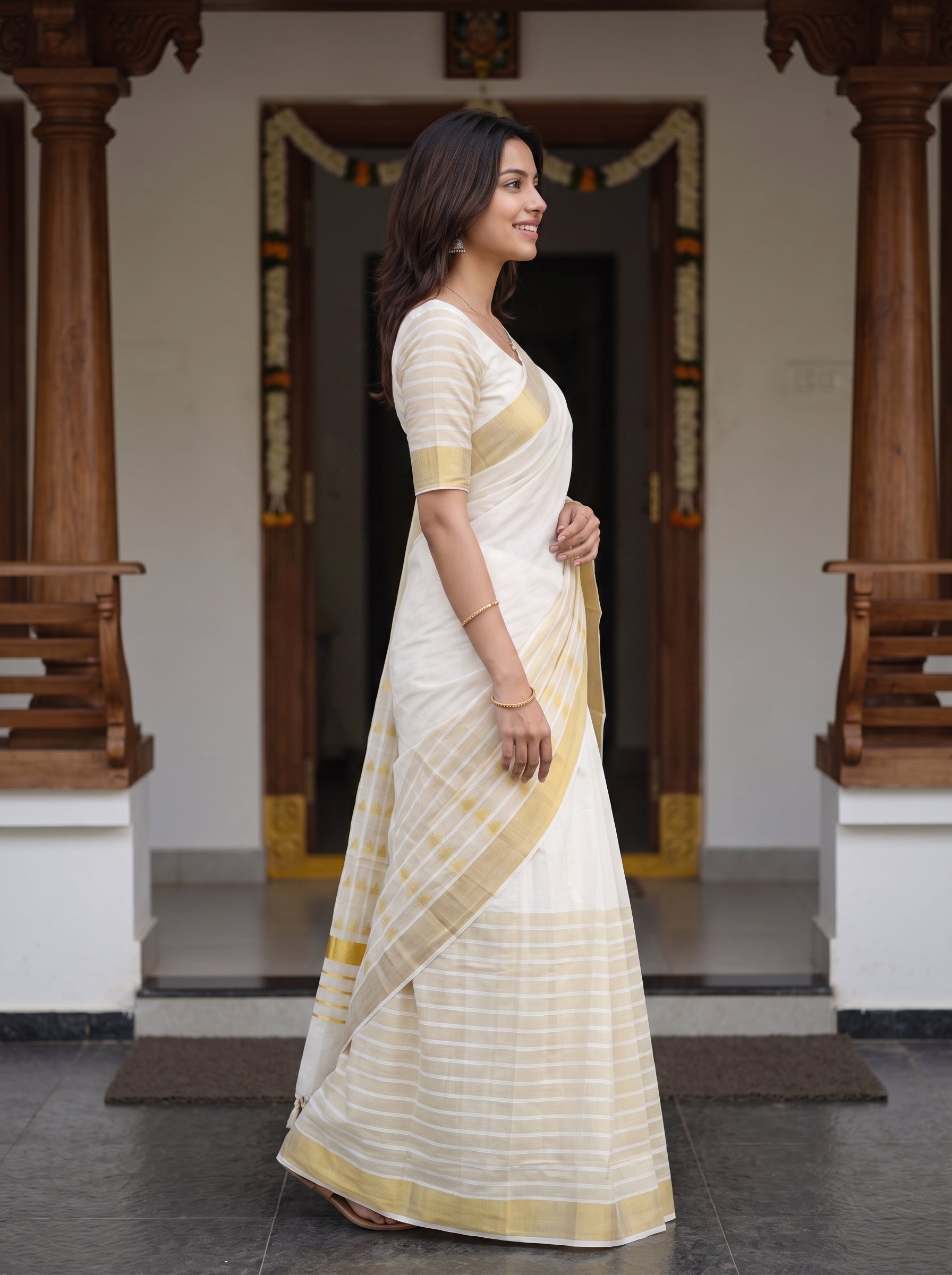 A woman wearing a white and gold striped saree stands in profile in front of a traditional doorway with wooden pillars.