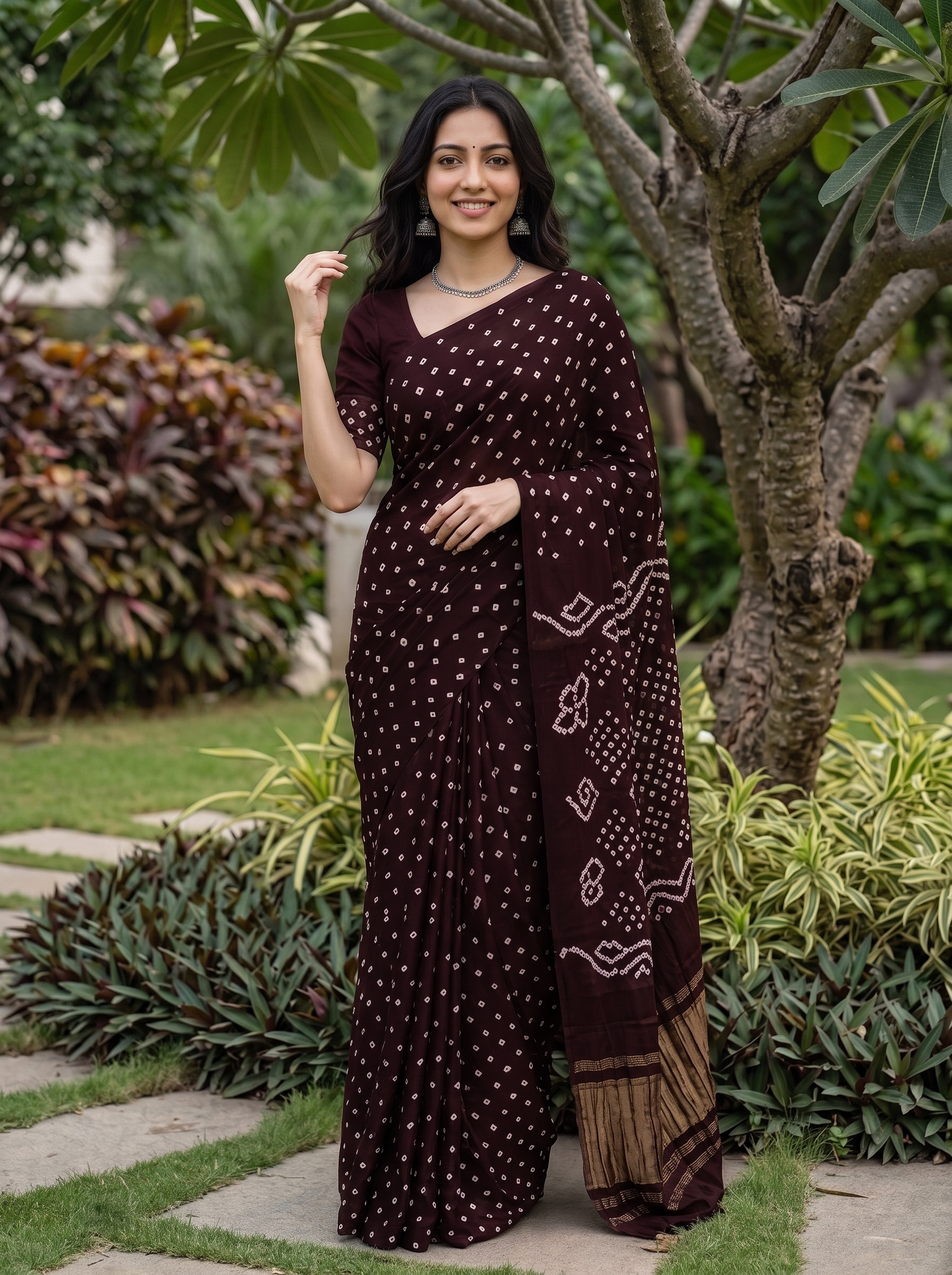 A woman stands outdoors in a garden wearing a dark maroon/burgundy saree with small white tie-dye dots and a patterned border. The background features green grass, plants, and trees.