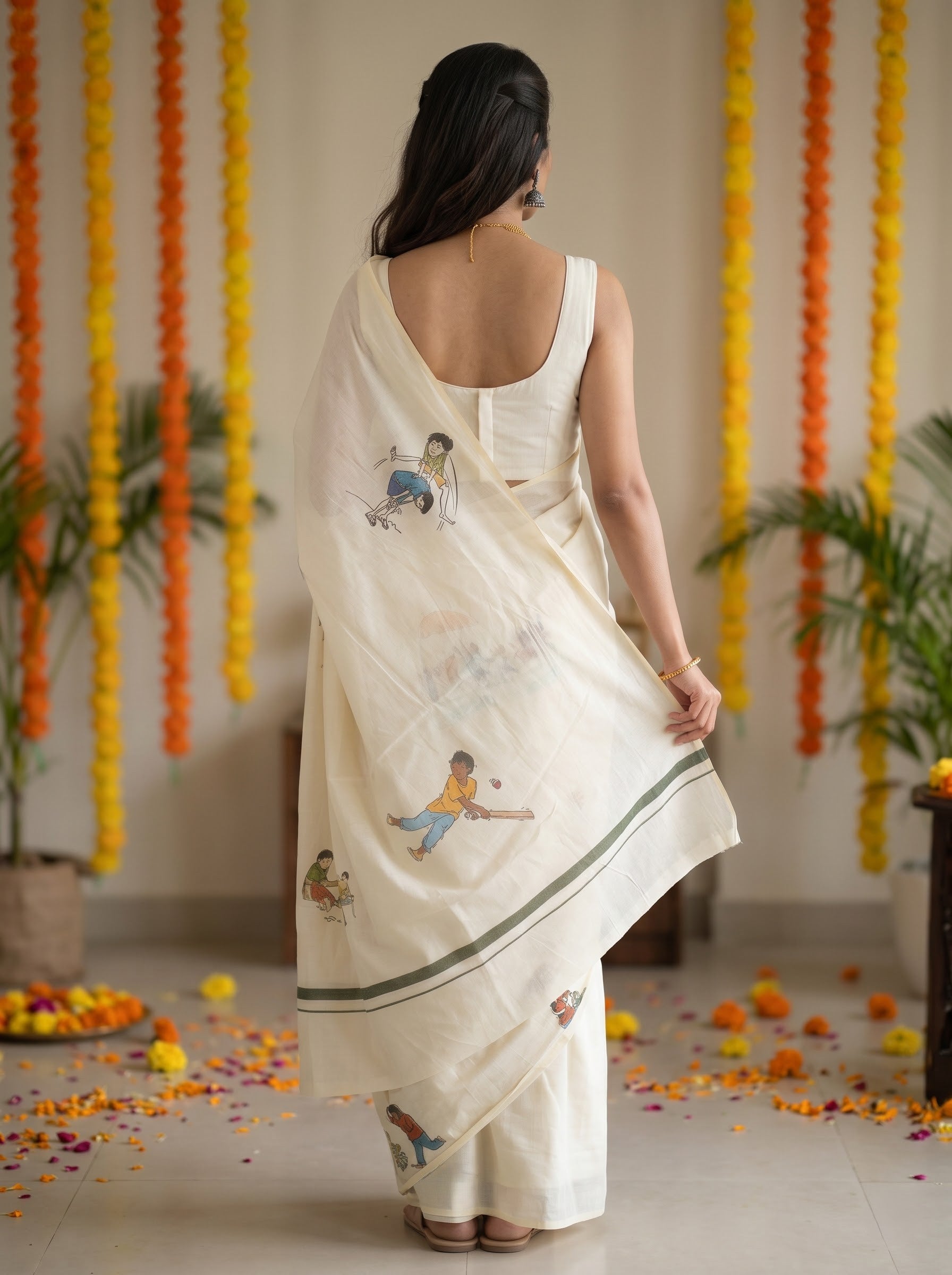 A woman viewed from behind wearing a cream-colored cotton saree with hand-painted folk art motifs of children playing, set against a decorated indoor background with hanging marigold garlands.
