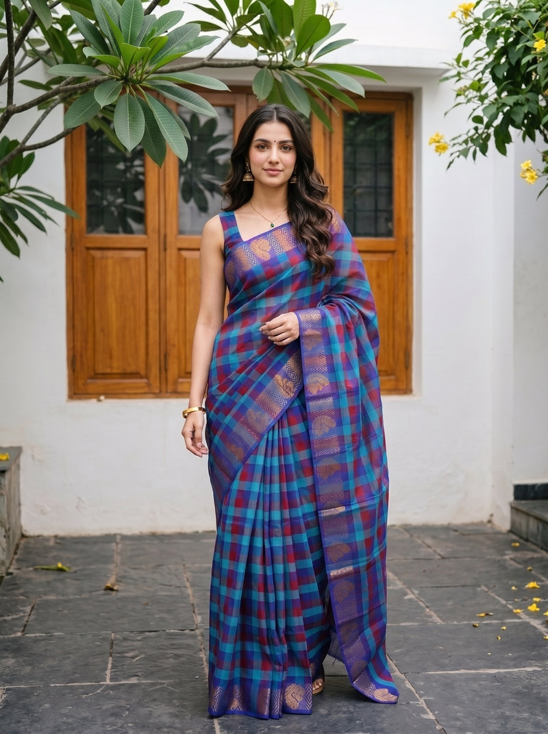 A woman wearing a purple and blue plaid silk saree with gold zari detailing, standing outdoors in front of a white wall with wooden doors.