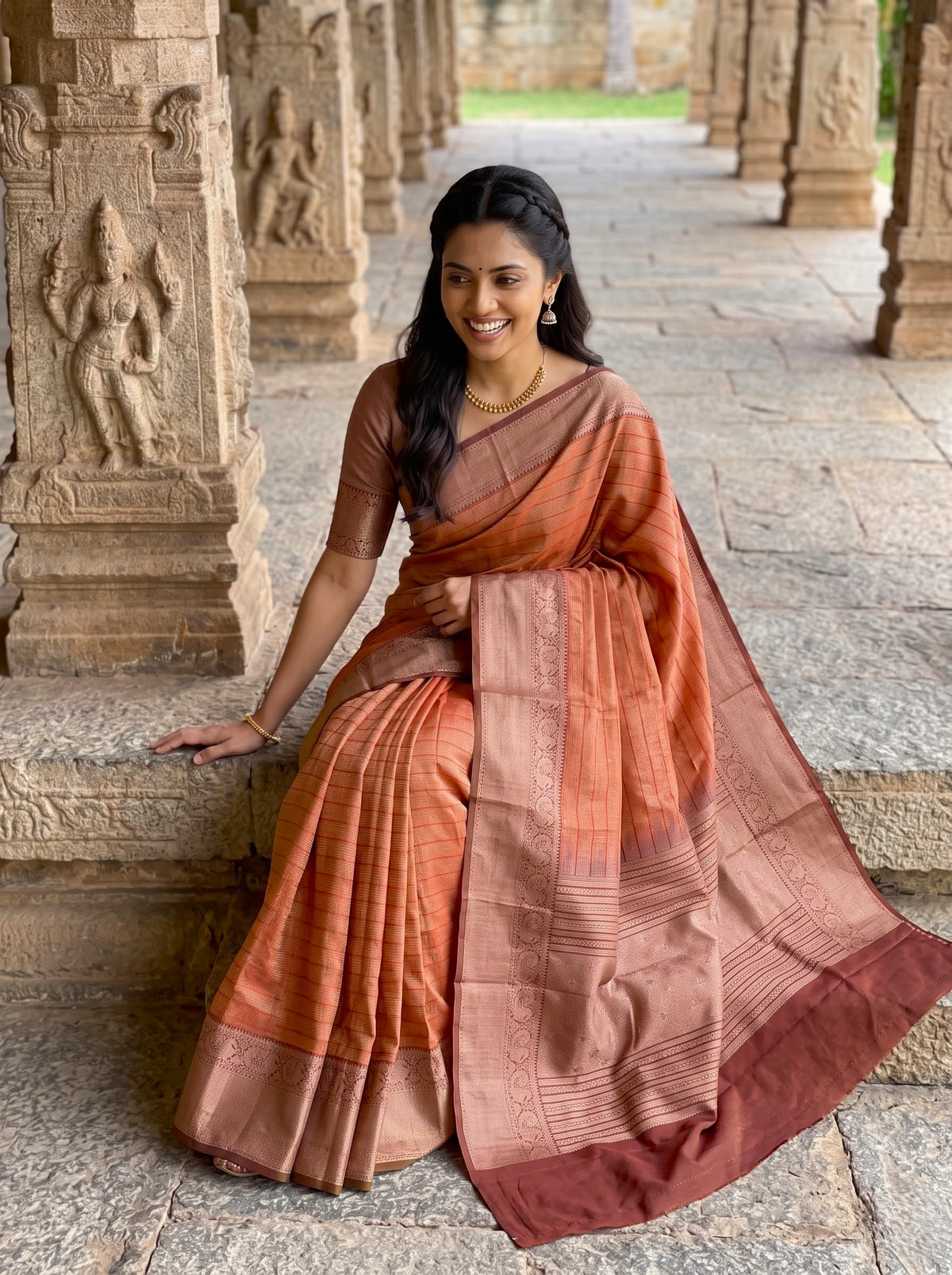 A woman wearing a rust-orange and terracotta silk saree with a metallic gold border, seated on stone steps in an ancient temple corridor.
