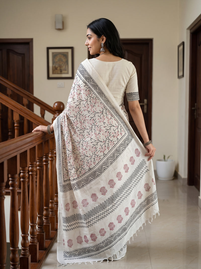 A woman wearing a cream-colored saree with pink floral prints and gray geometric borders, standing in a home hallway next to a wooden staircase.