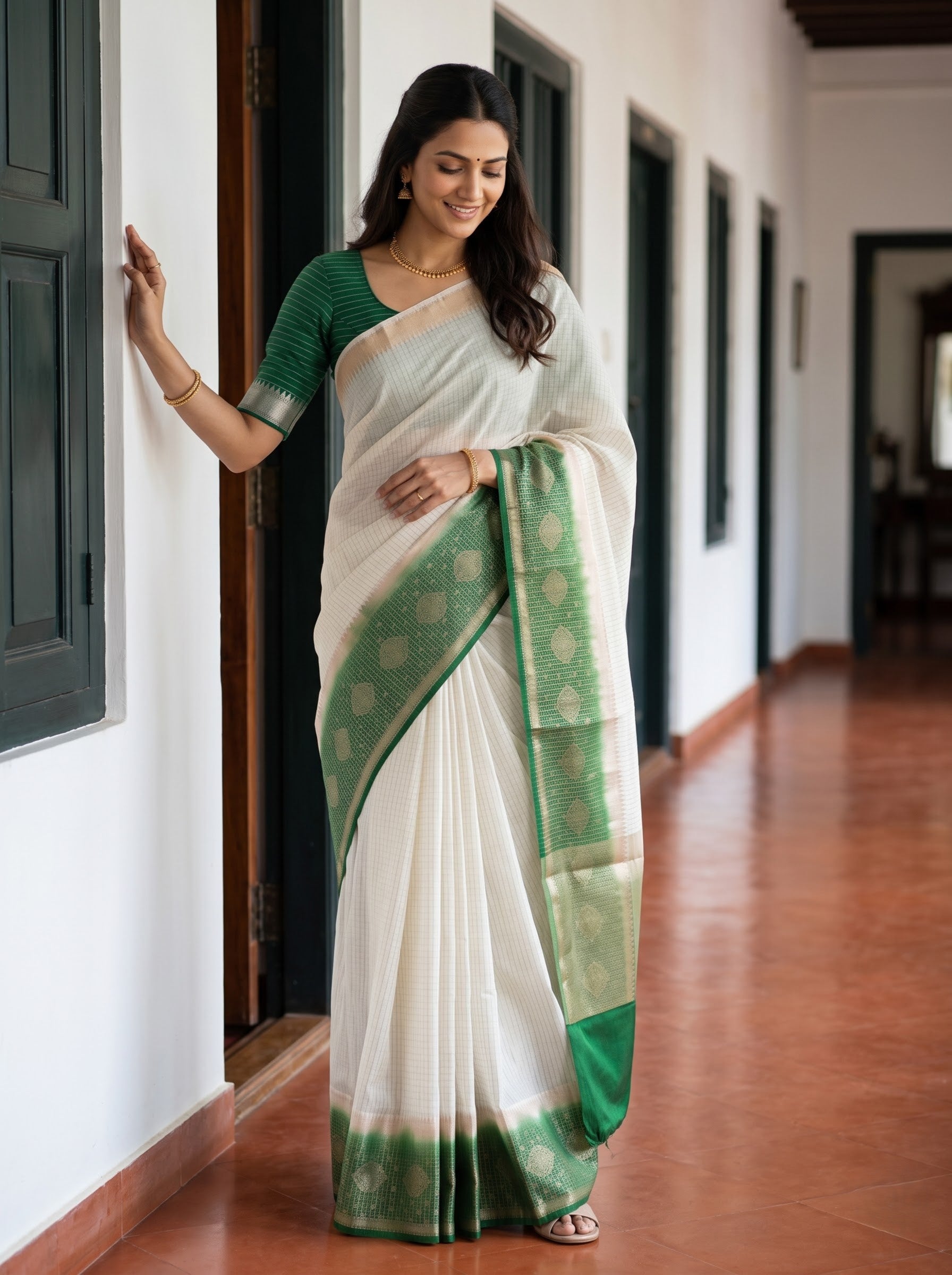 A woman wearing a white and green saree stands in a hallway with white walls and dark door frames, holding onto a wall for support.
