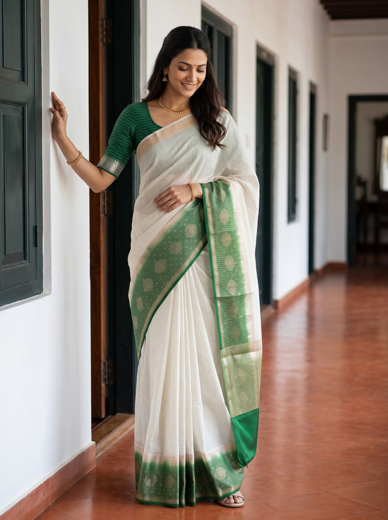 A woman wearing a white and green saree stands in a hallway with white walls and dark door frames, holding onto a wall for support.