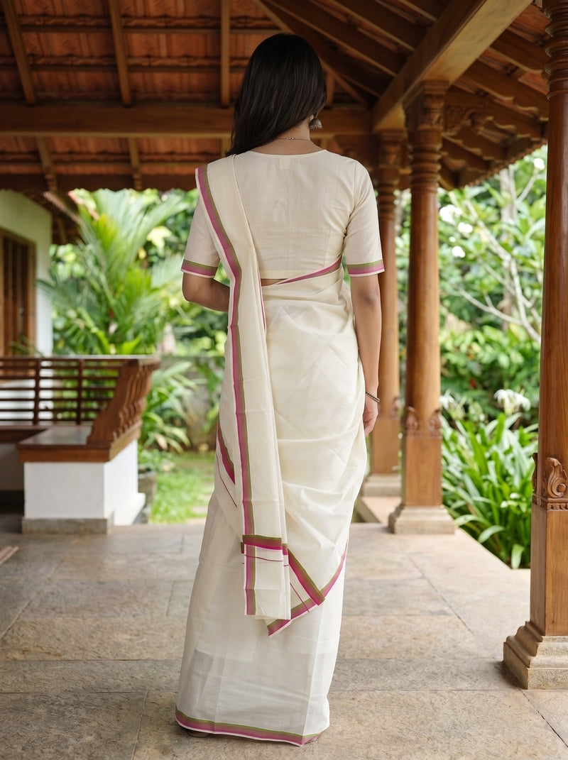 A woman viewed from behind wearing a cream-colored cotton saree with pink border accents, standing on a wooden porch with lush green garden foliage in the background.