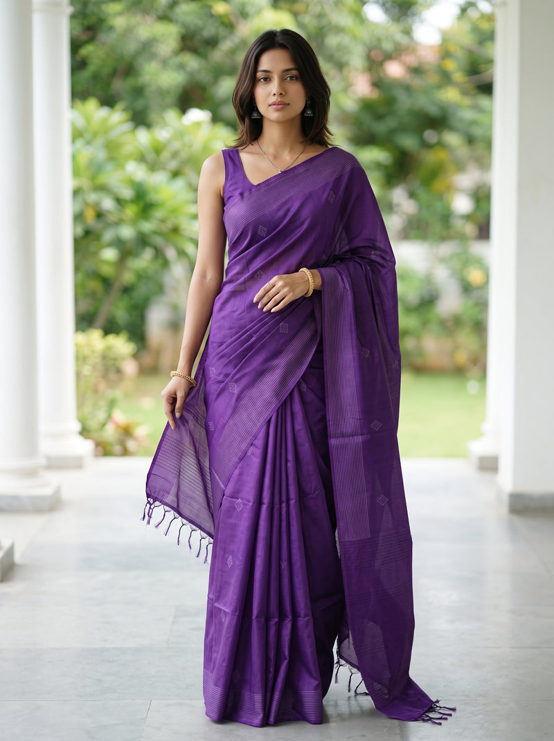 A woman modeling a purple silk saree with a matching sleeveless blouse, standing in a covered walkway with green garden foliage in the background.