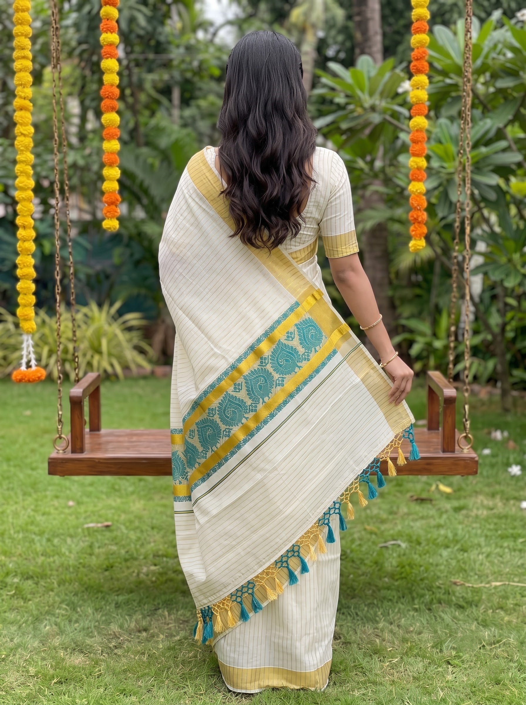 A woman viewed from behind wearing a cream-colored saree with yellow and teal borders, standing on a green lawn in front of hanging wooden swings decorated with marigold garlands.