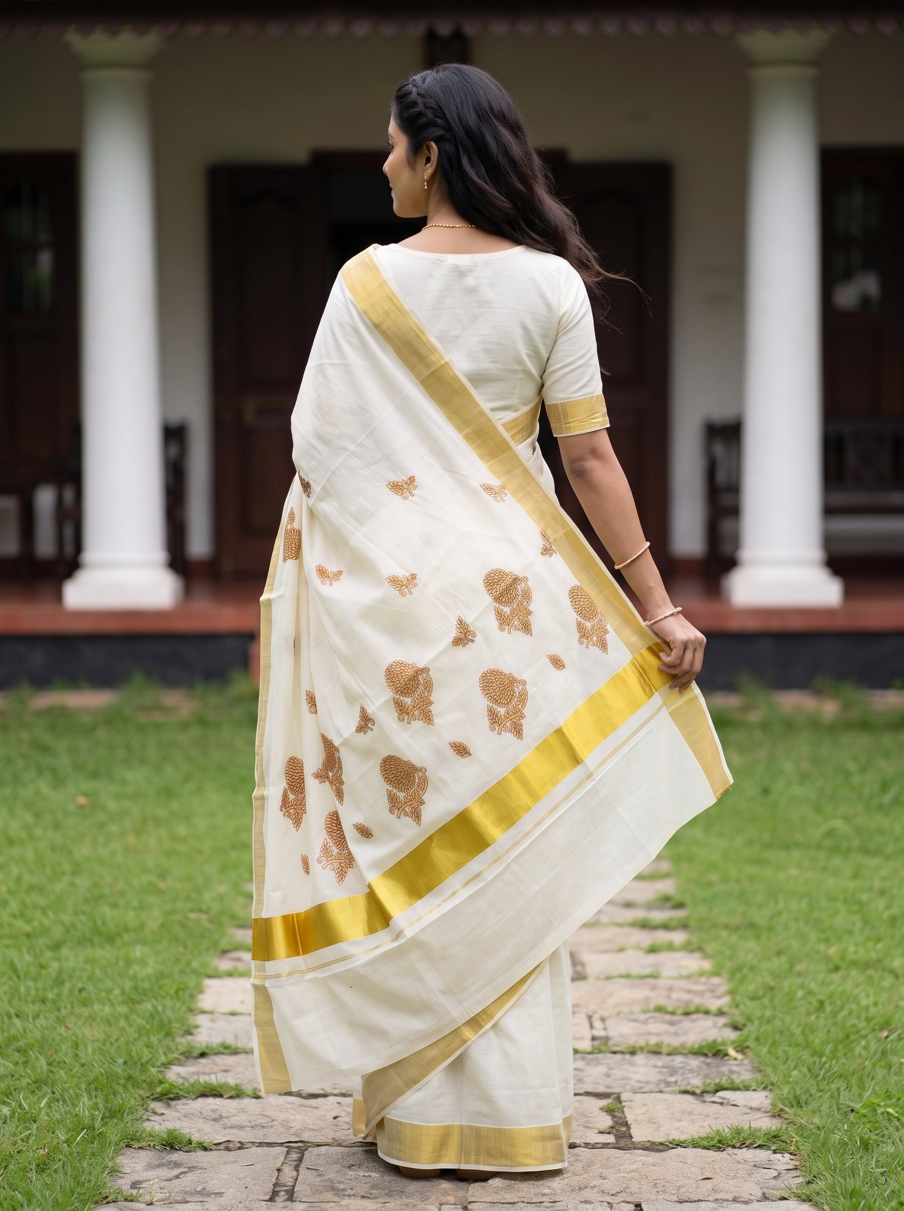 A woman wearing a cream-colored saree with gold borders and brown floral motifs, standing on a stone path in front of a building with white pillars.